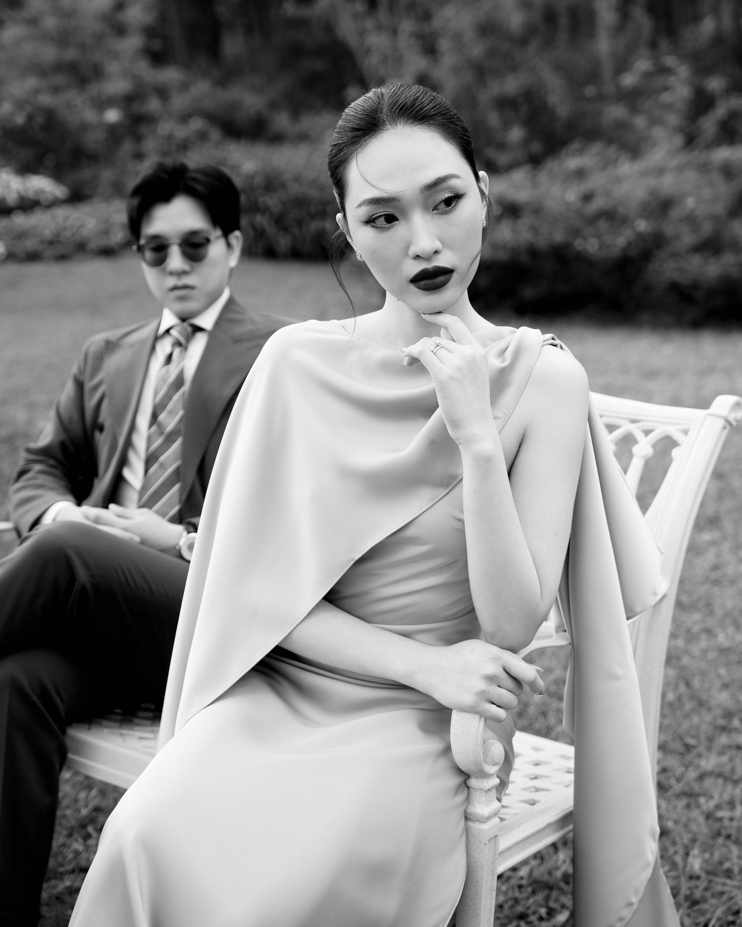 Black and white photo of stylish couple in formal attire seated in garden.