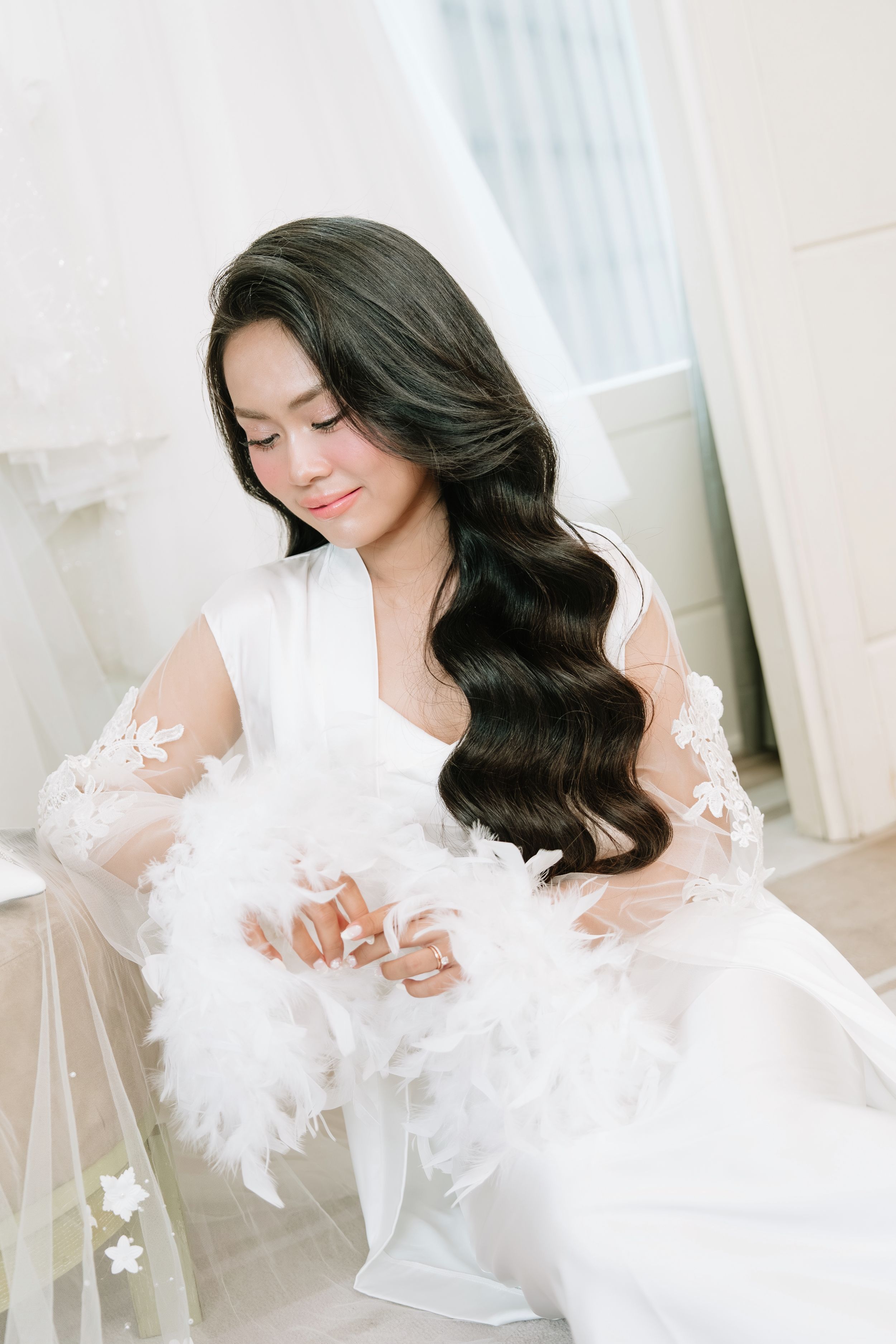 Beautiful bride with long wavy hair in white lace robe holding feather bouquet in a bright bridal suite.