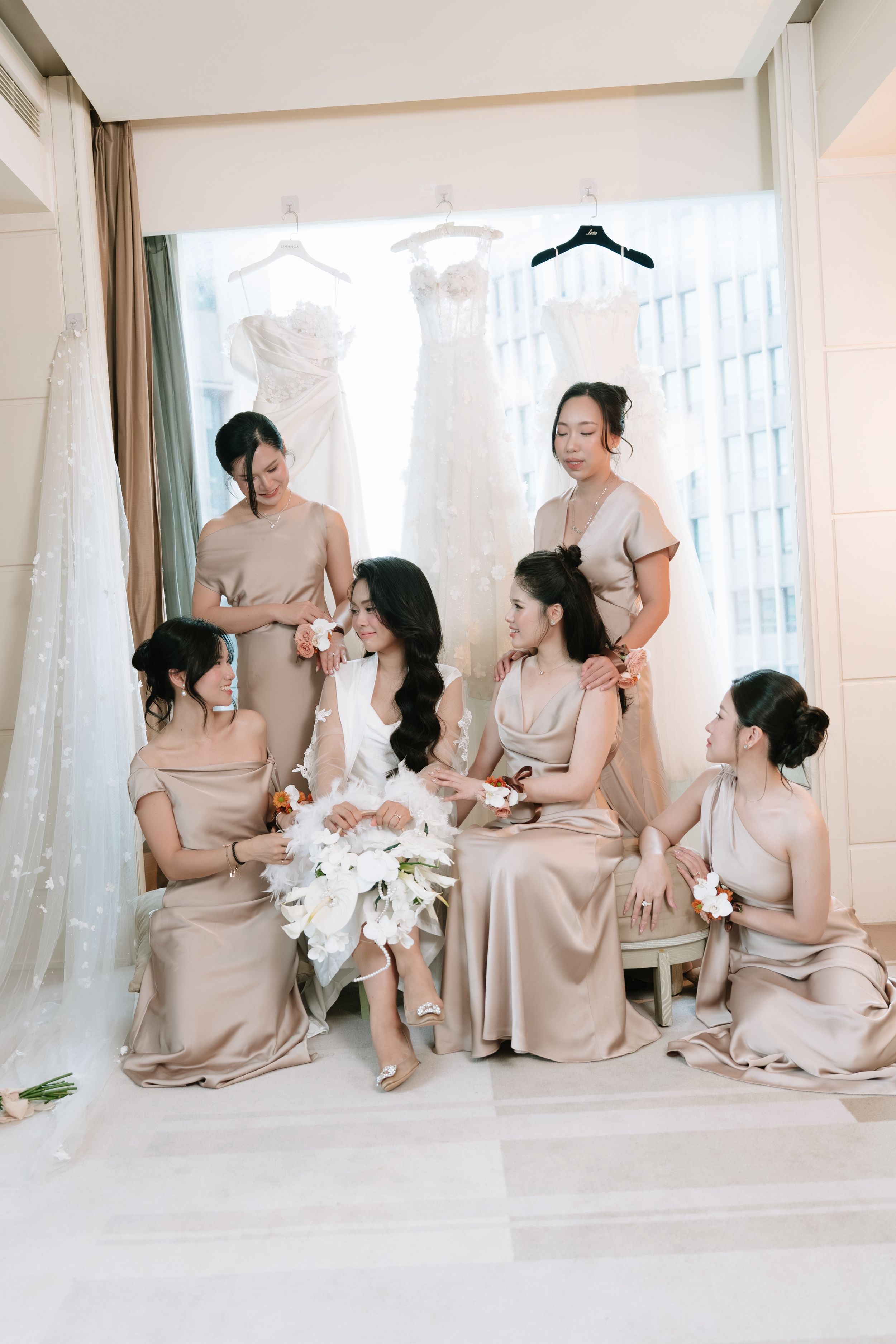 Bride in white robe surrounded by six bridesmaids in champagne satin dresses with wedding gown backdrop.