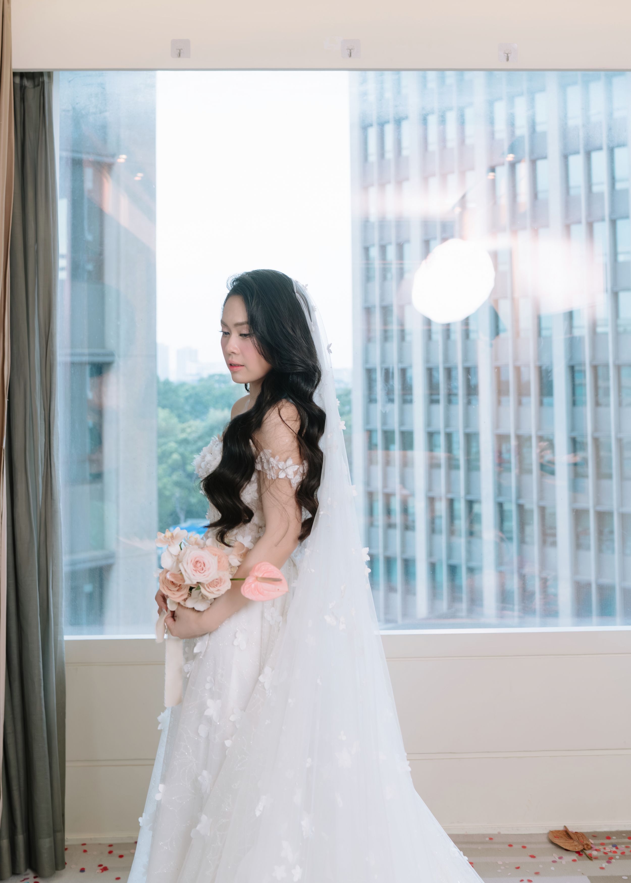 Bride in ornate white wedding gown with veil holding bouquet, standing by large windows in soft natural light.