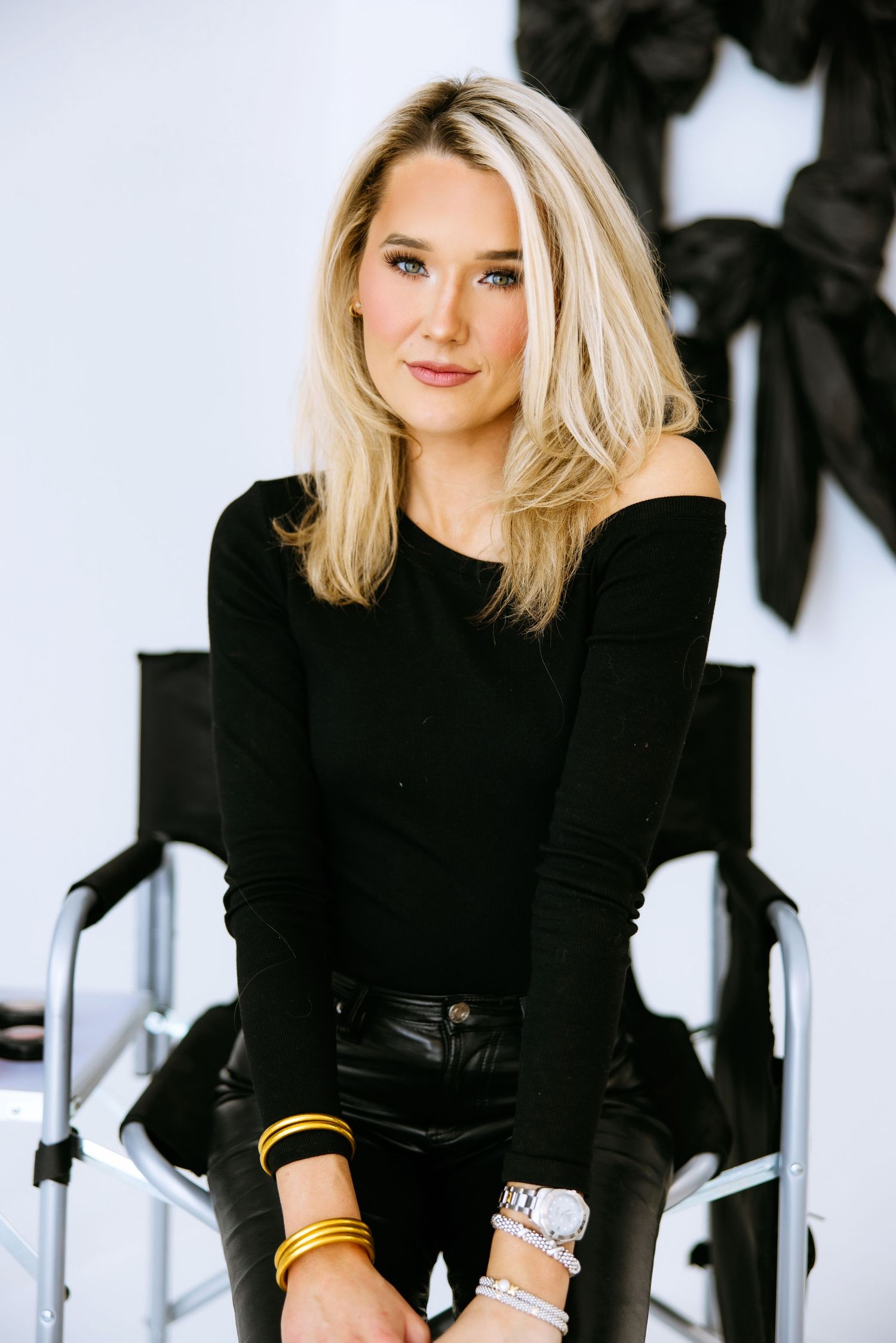 A stylish fashion portrait featuring a blonde model in a black sweater and leather pants against a white backdrop.
