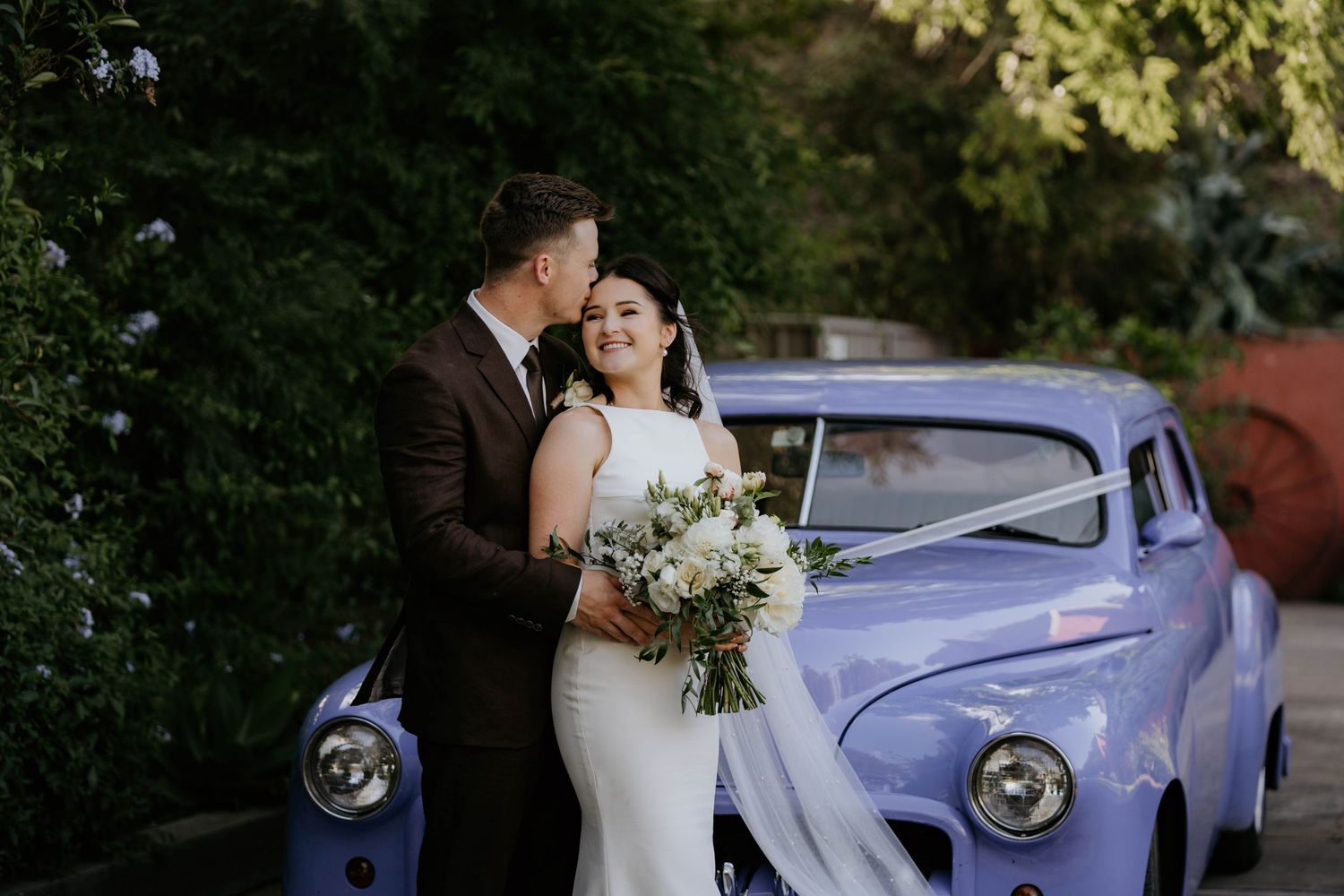 Newlywed couple embraces beside a vintage light blue classic car holding a white floral bouquet.