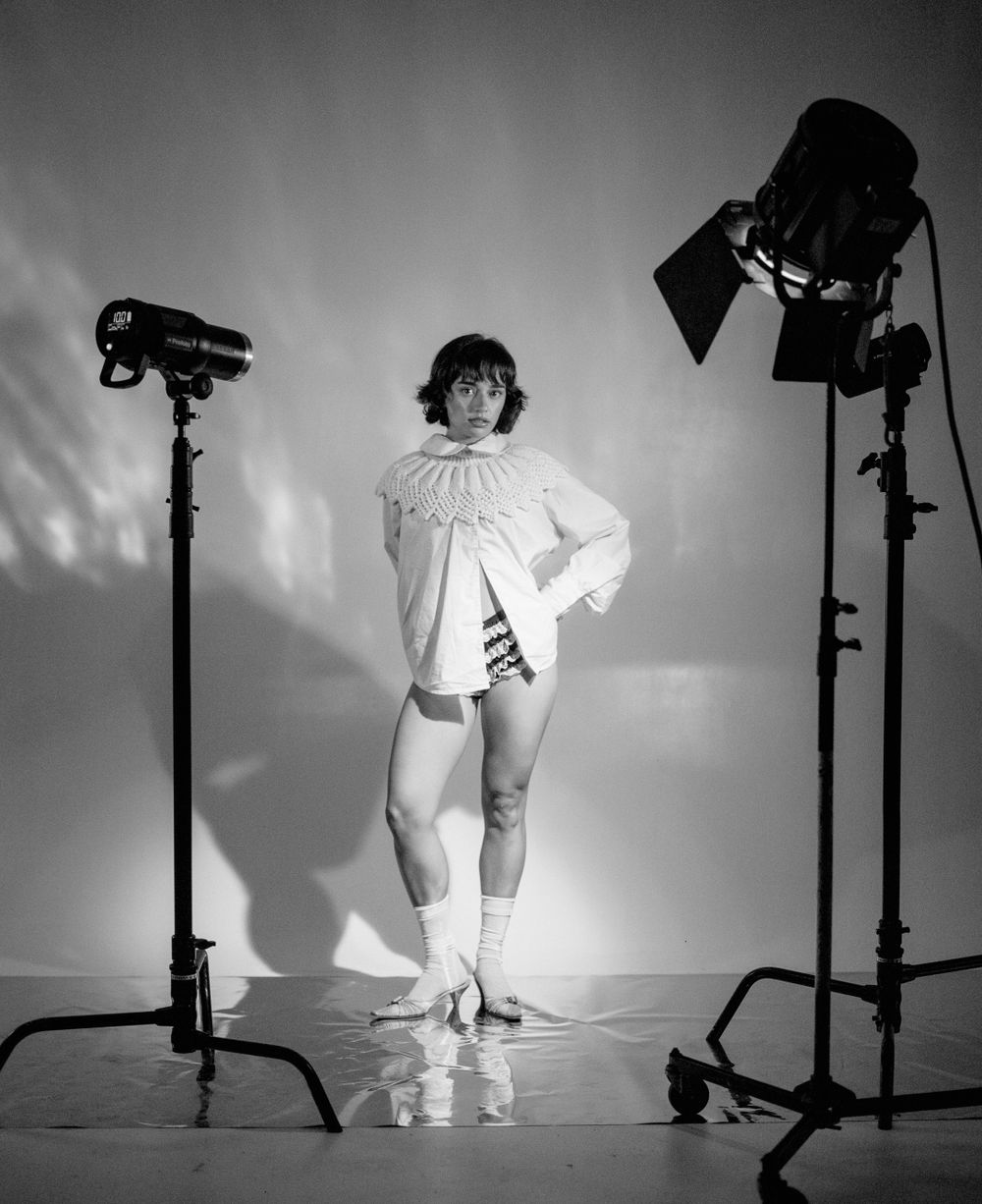 Studio photography setup with dramatic lighting showing figure in white ruffled collar and knee socks against light backdrop.