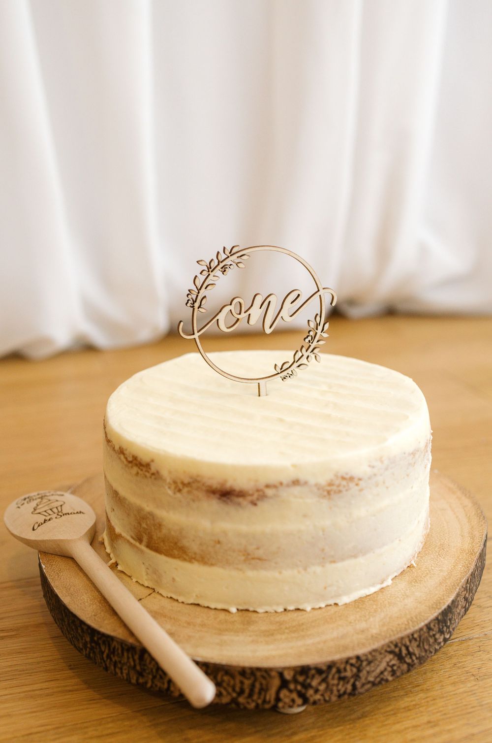 A naked frosted first birthday cake with a wooden 'one' topper on a rustic wood slice with a decorative spoon.