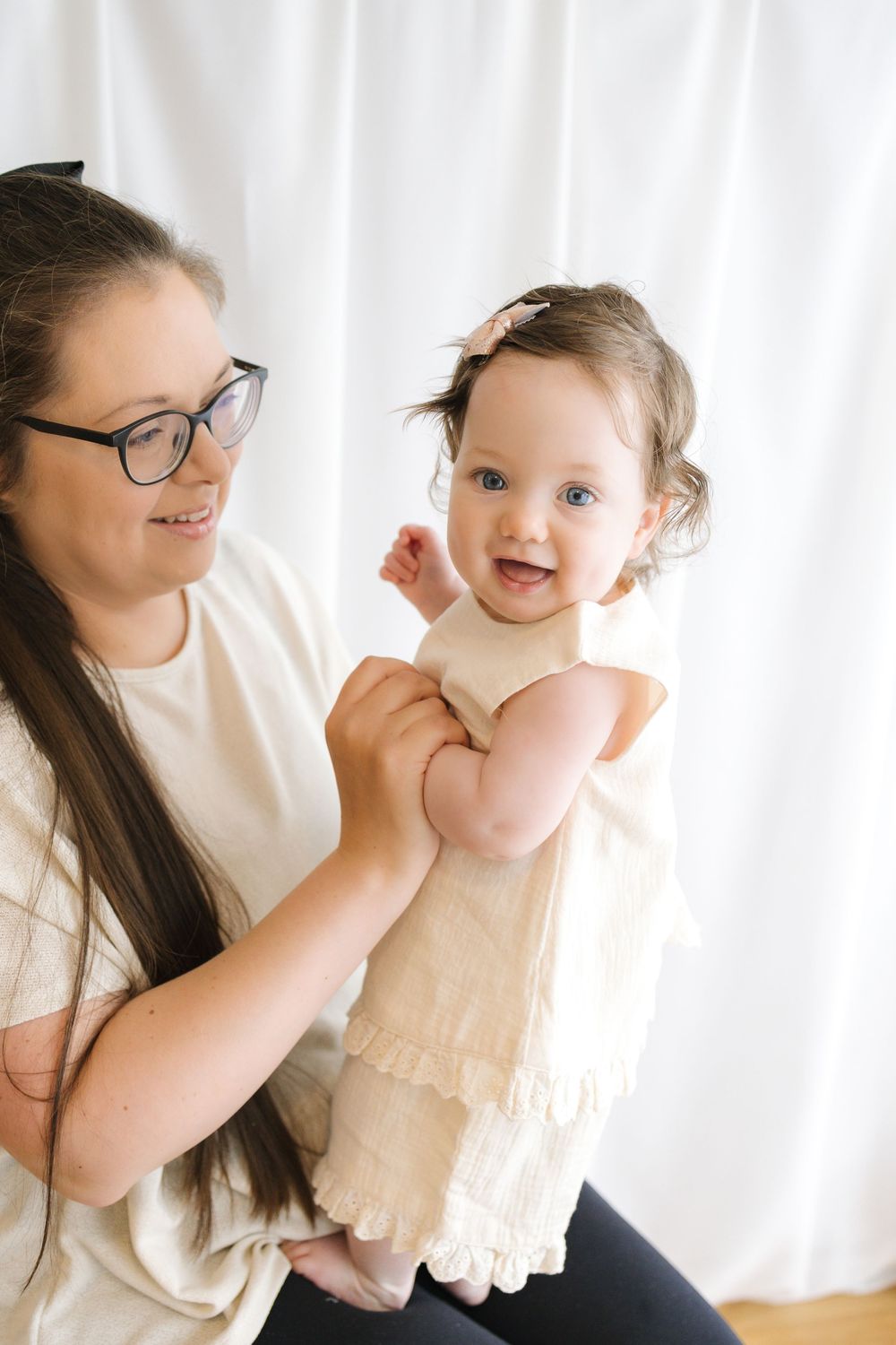 A woman with glasses holds a smiling baby girl in a cream dress with a bow headband against a white backdrop.