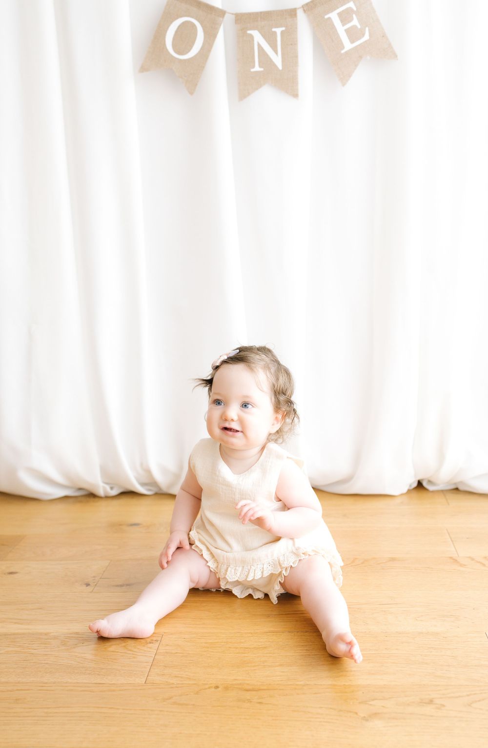 Baby girl in cream romper sits on wood floor smiling, with burlap ONE banner for first birthday photo.