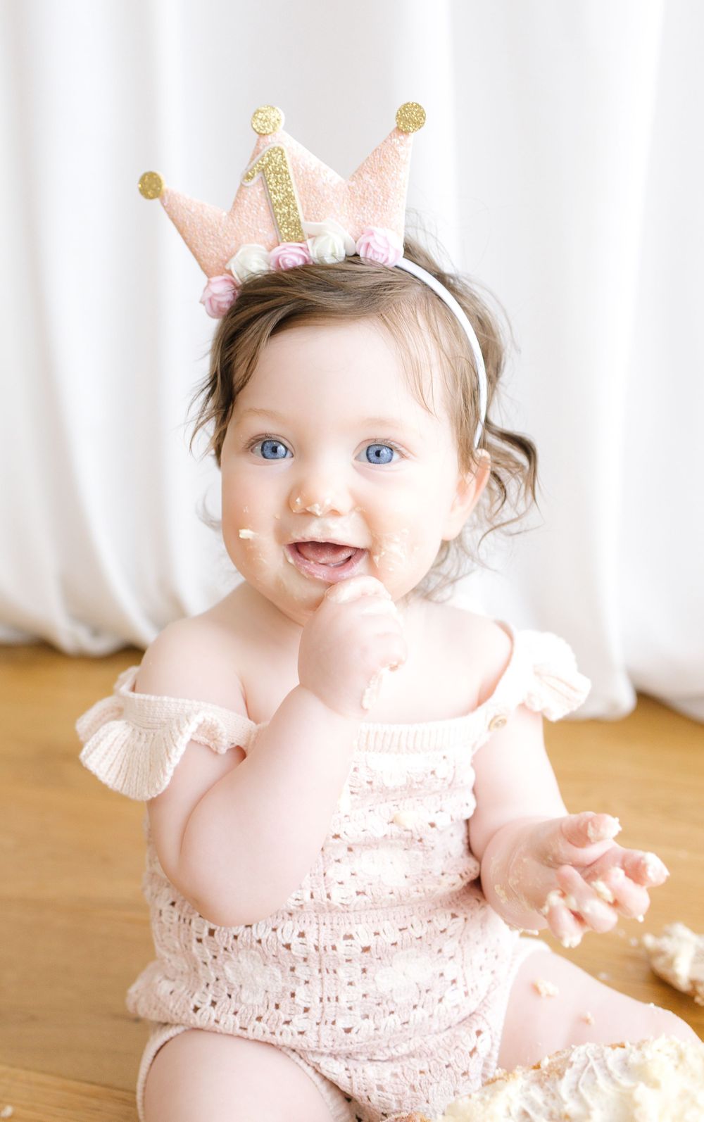 A blue-eyed baby girl in a pink romper and birthday crown enjoys her first cake smash celebration.