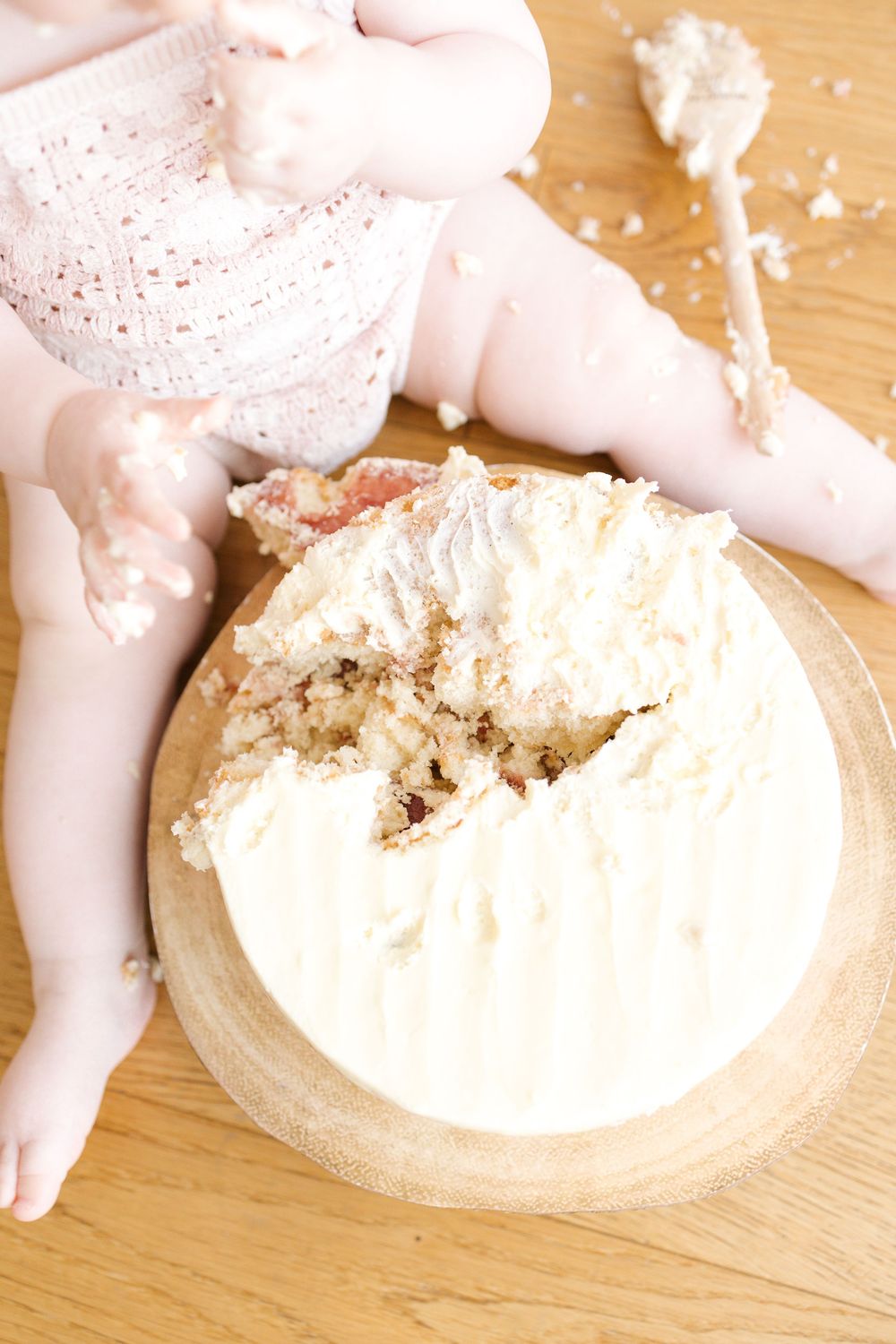 Baby's first birthday cake smash with white frosted cake on wooden board, partially eaten on hardwood floor.