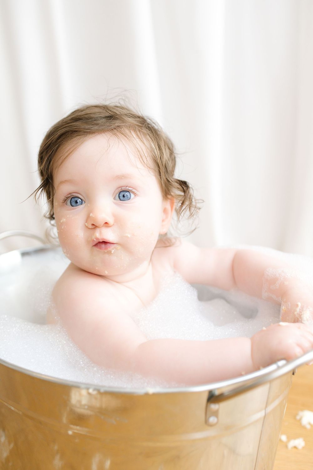 A blue-eyed baby sitting in a metal tub filled with soapy bubbles, looking curiously at the camera.