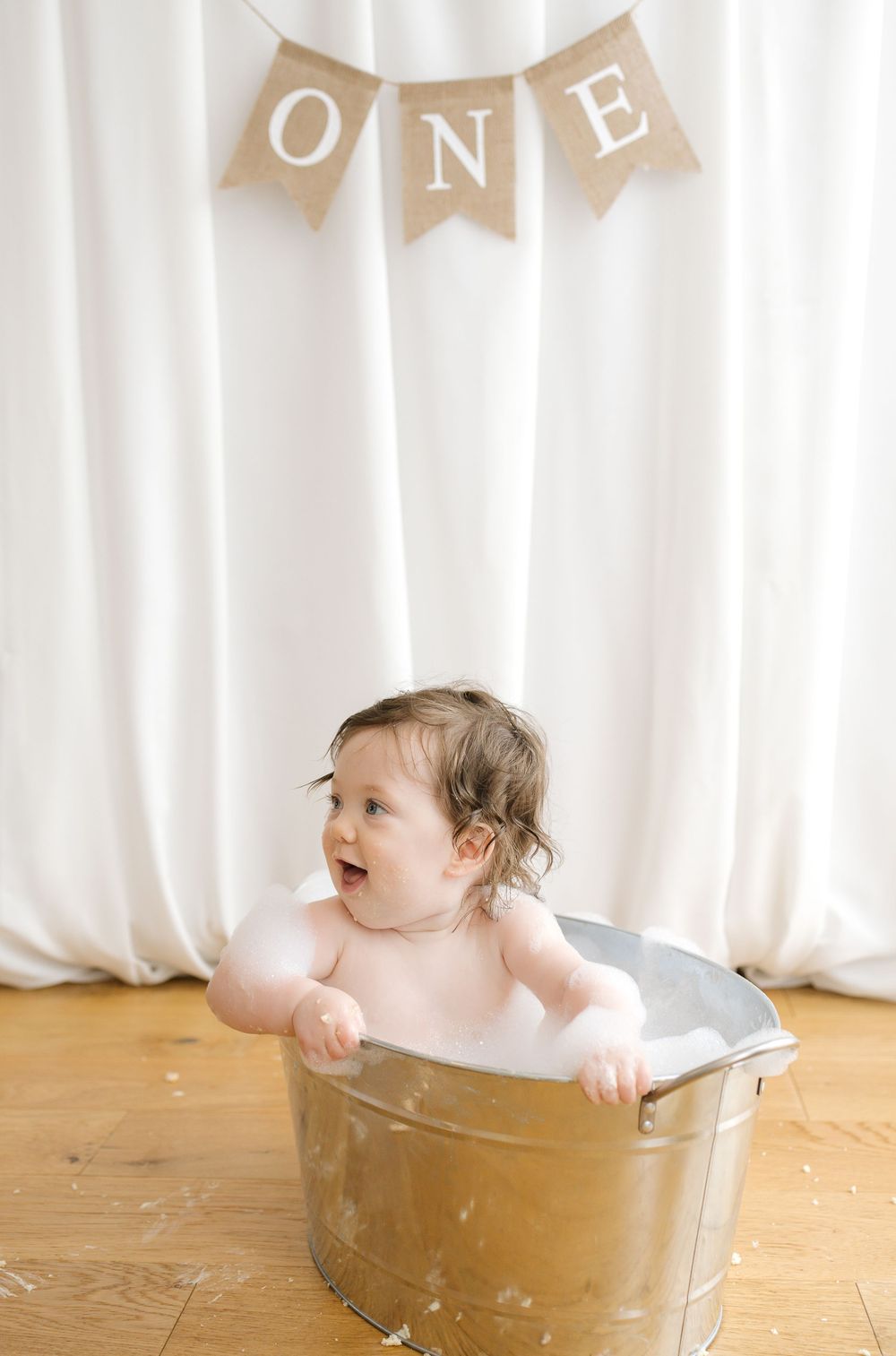 A happy baby sits in a galvanized tub during a first birthday milestone bath photo session with an ONE banner.