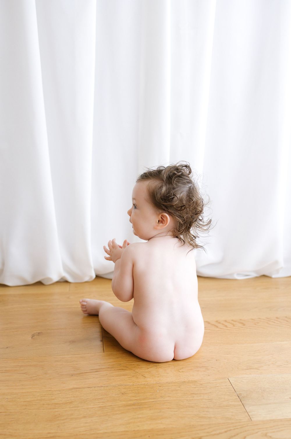 Naked baby with curly hair sitting on hardwood floor facing white curtains.