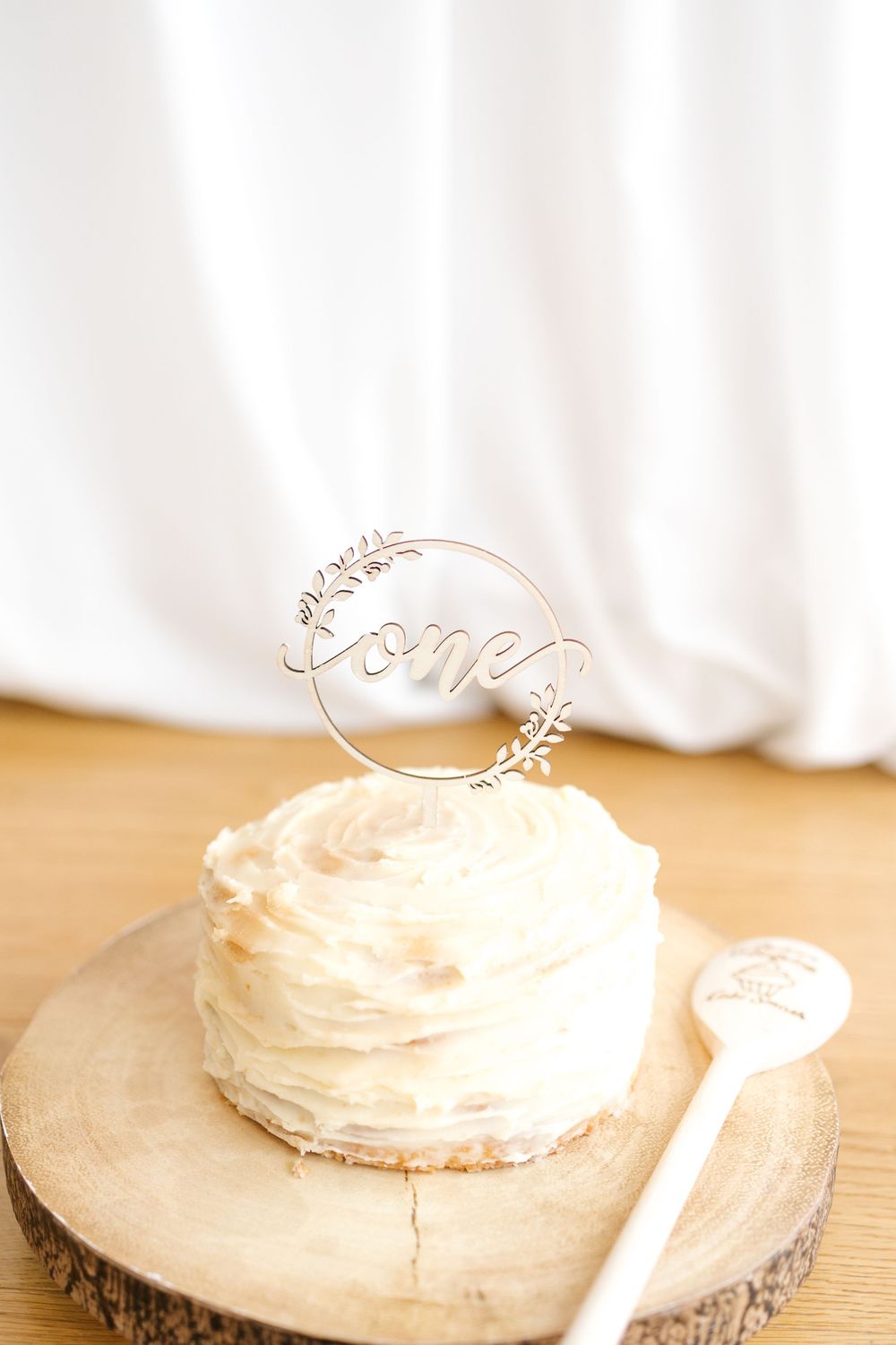 A white frosted first birthday smash cake with a gold \"one\" topper sits on a rustic wood slice with a decorative spoon.