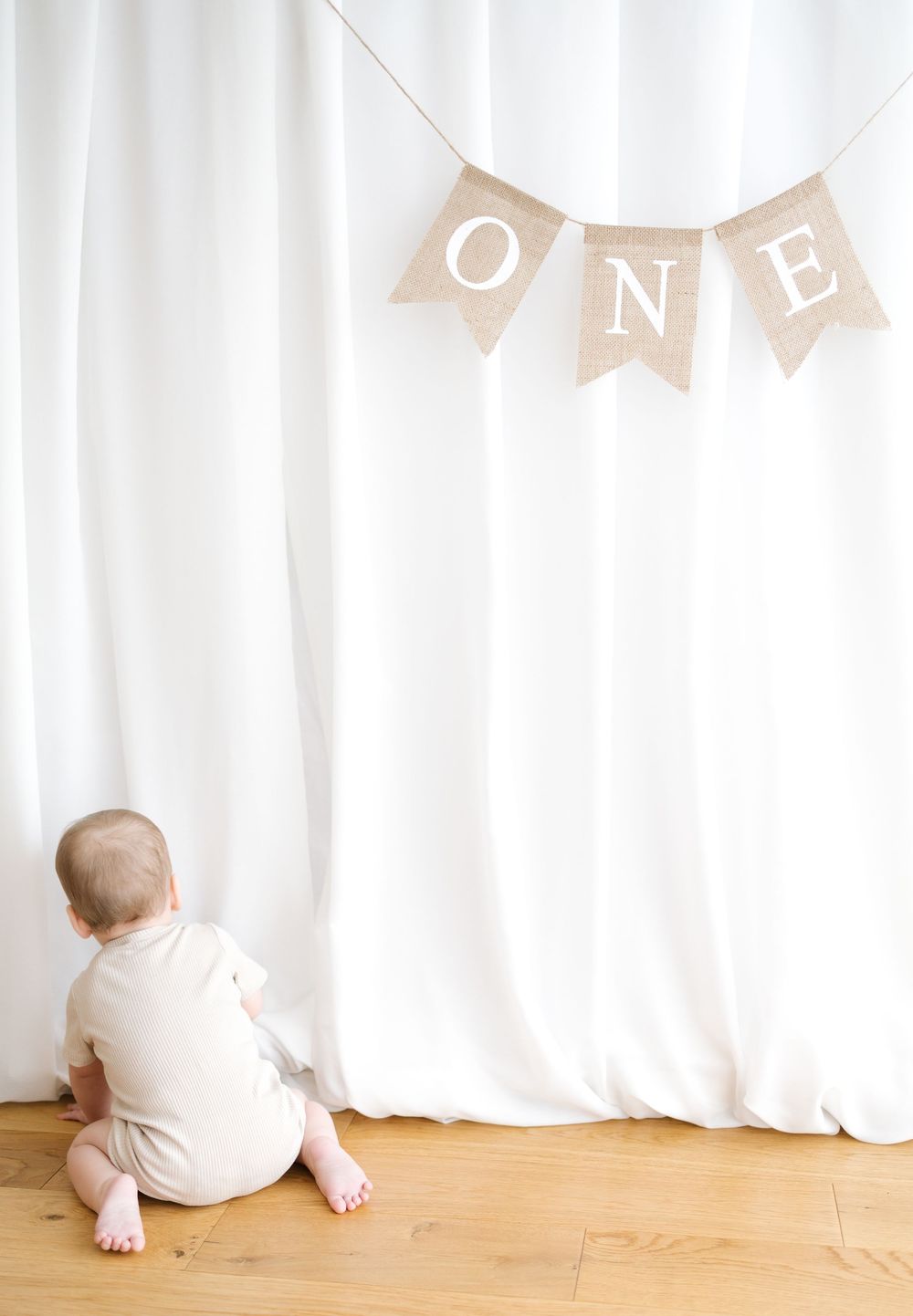 Baby sitting on wood floor facing white curtain backdrop with burlap ONE banner above for first birthday.