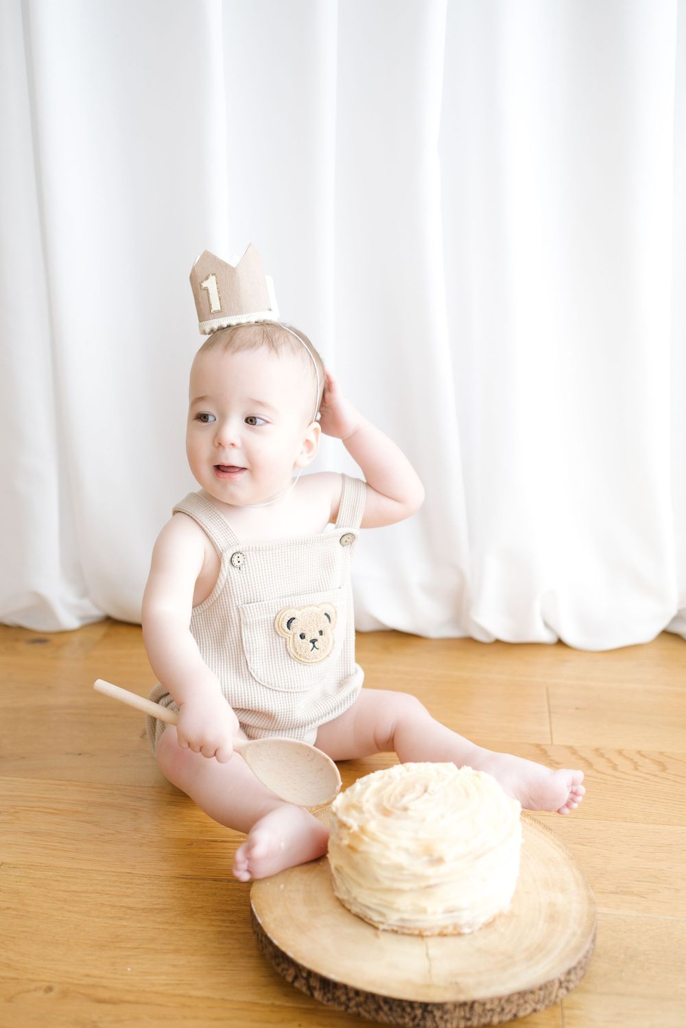 Baby in bear romper and crown sits with smash cake and wooden spoon celebrating first birthday.