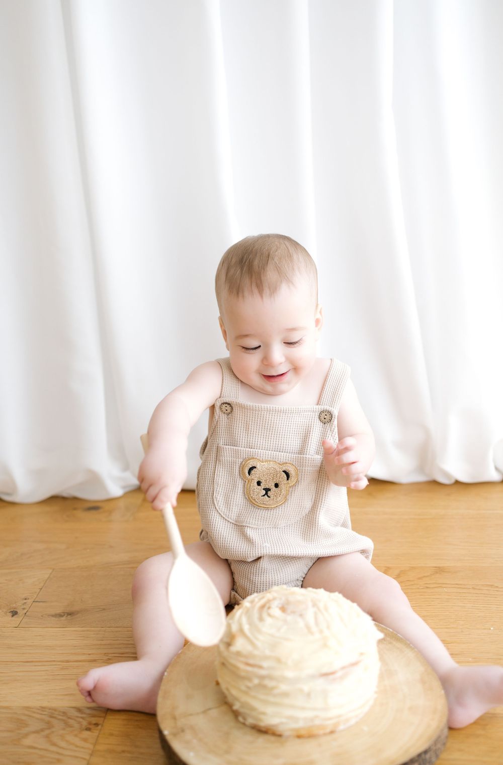 Smiling baby in bear overalls sits on wood floor with spoon and frosted cake for first birthday smash.