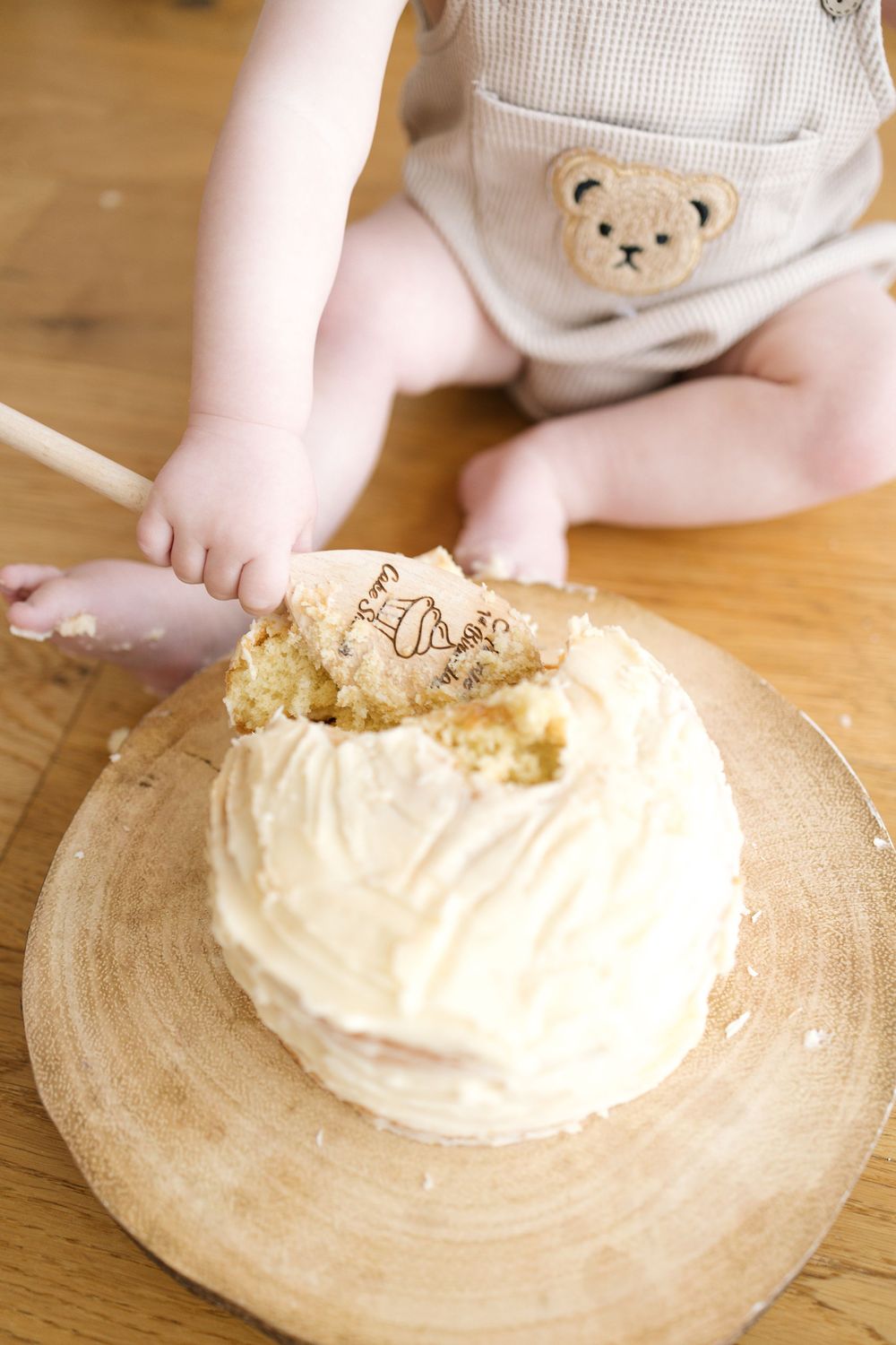 Baby in bear overalls smashing a white frosted smash cake on a wooden board for first birthday.