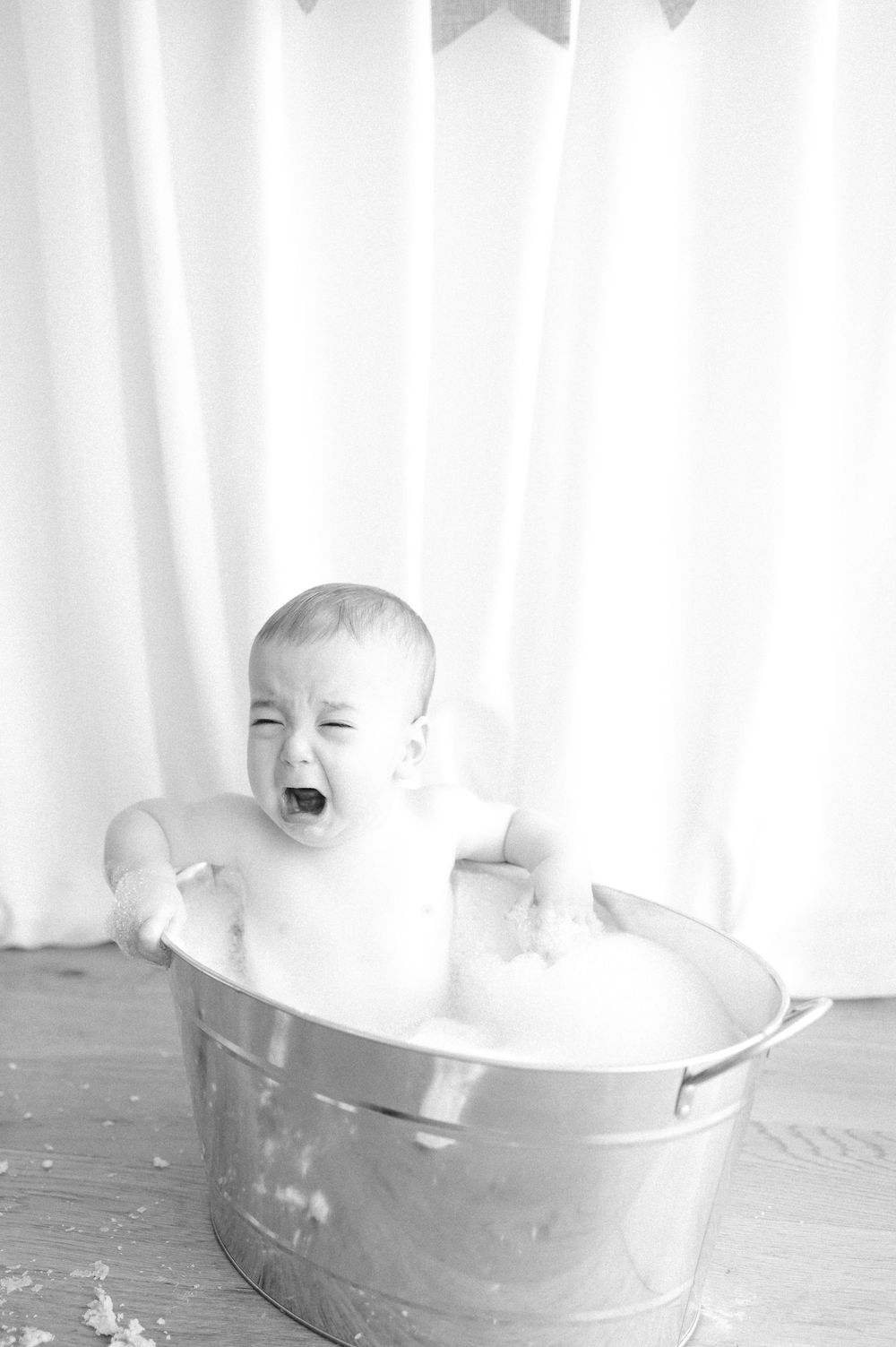 Black and white photo of a crying baby sitting in a metal tub filled with bubbles during bath time.