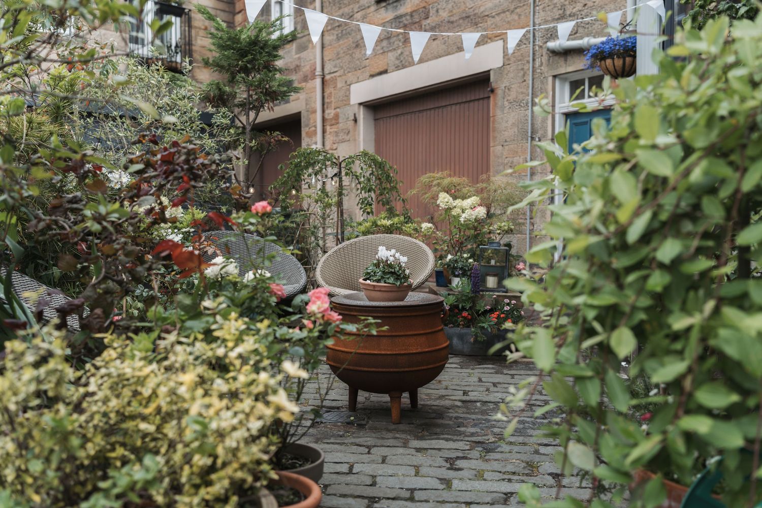 A rustic patio garden with a metal fire pit, decorative arch, and brick wall surrounded by lush plants and potted flowers.
