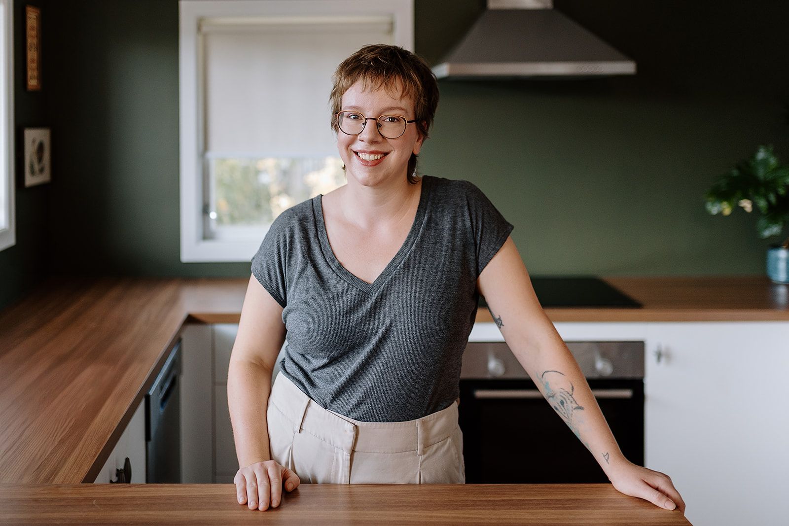 A person wearing a gray v-neck shirt and glasses stands smiling in a modern kitchen with wood countertops and green walls.
