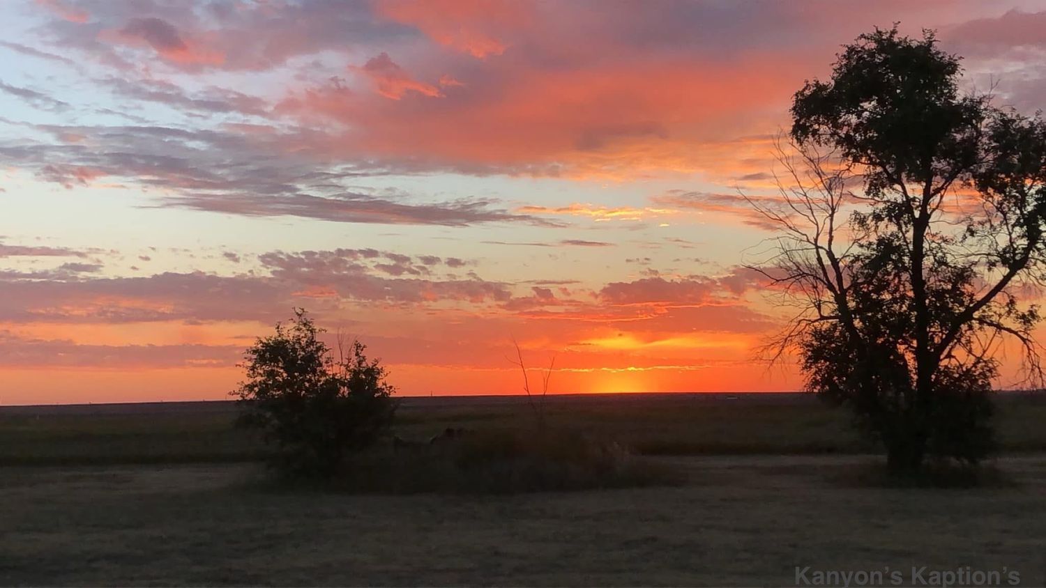 Dramatic orange and pink sunset over a dark rural landscape with silhouetted trees against the colorful sky.