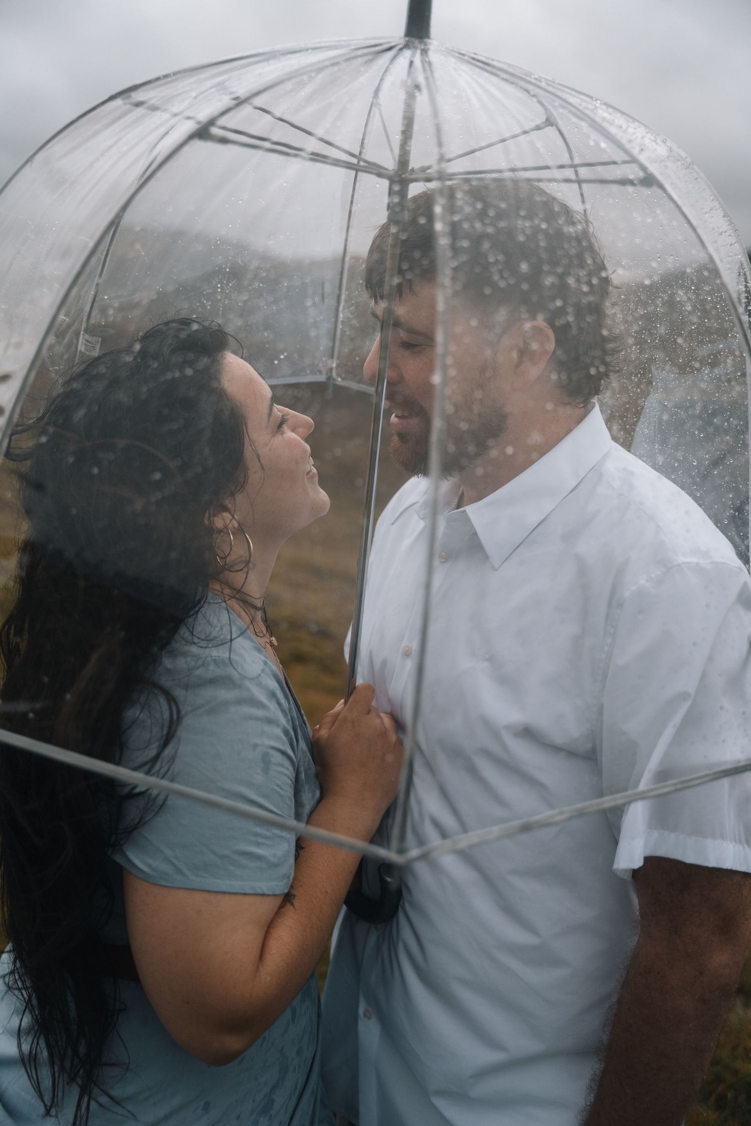 A couple shares an intimate moment under a clear umbrella in the rain.
