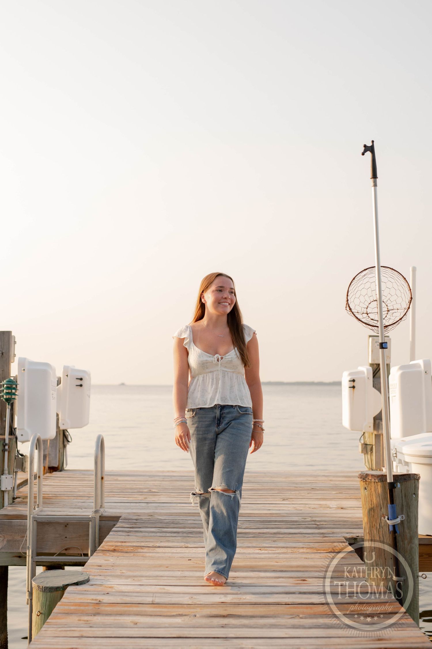 Person in white top and distressed jeans walking on marina dock at sunset.