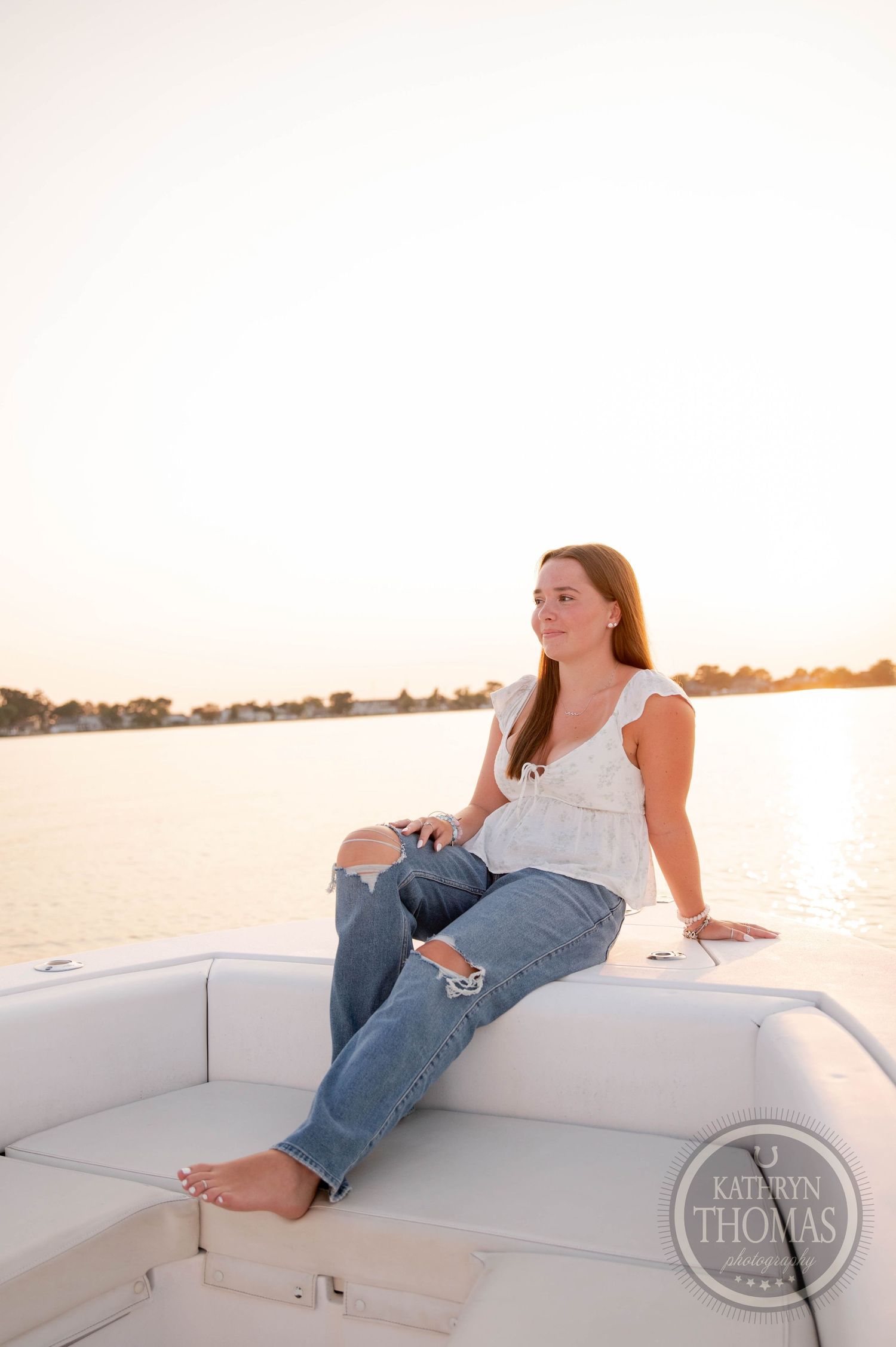 Woman in distressed jeans and white top sits on boat cushion during sunset.