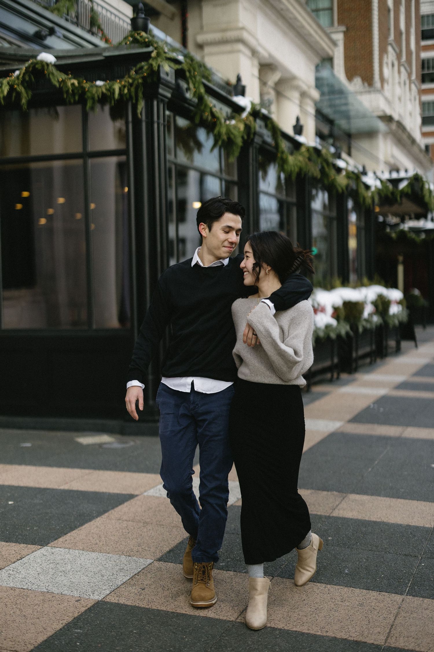 A couple enjoys a romantic winter moment walking together outside a decorated storefront with holiday greenery decor.