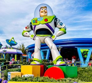 A large Buzz Lightyear statue stands on colorful blocks at a Disney theme park against a bright blue sky.