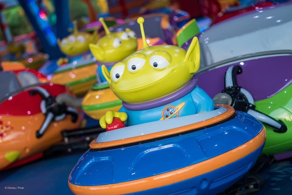 A green three-eyed alien toy figure from Toy Story rides in a colorful spinning amusement park ride.