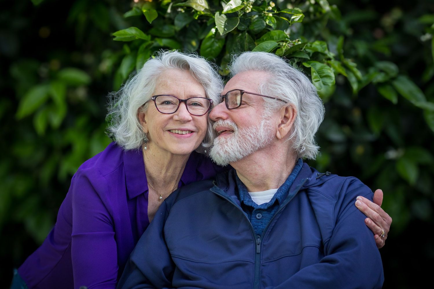 An affectionate elderly couple in glasses sharing a tender moment outdoors against green foliage.