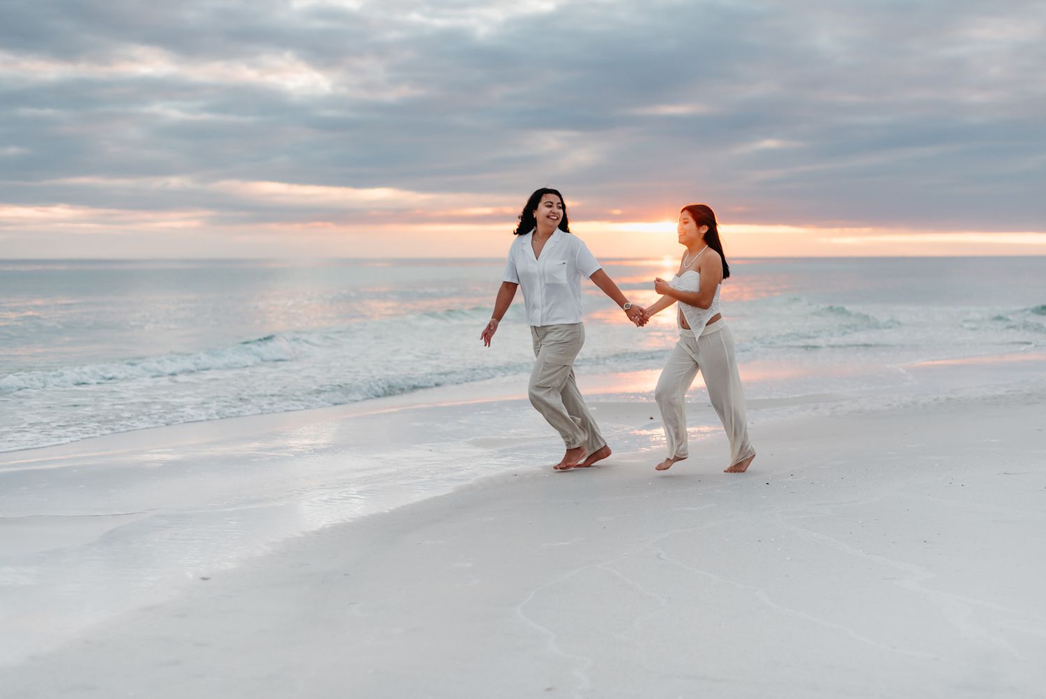Two people run joyfully along the beach shore at sunset wearing white outfits.