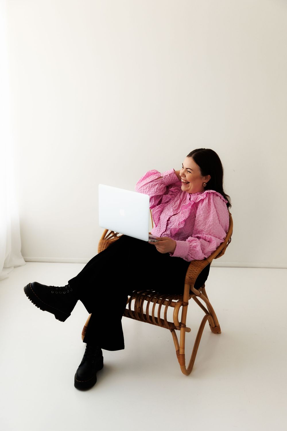 Woman in pink puff sleeve blouse and black pants laughing while reading documents in a wooden chair against white background.
