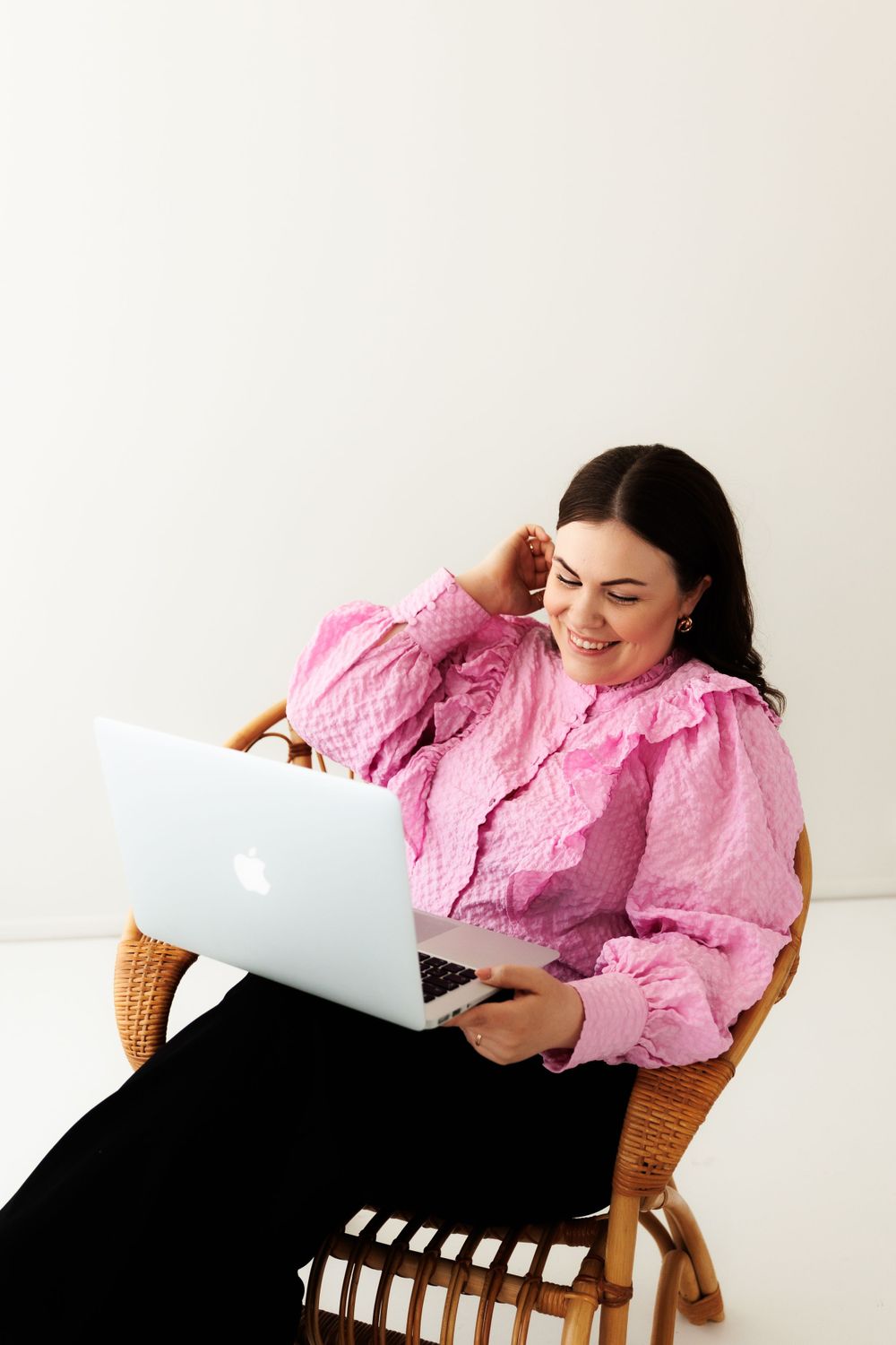 Woman in pink ruffle blouse sits comfortably in wicker chair while working on MacBook laptop.
