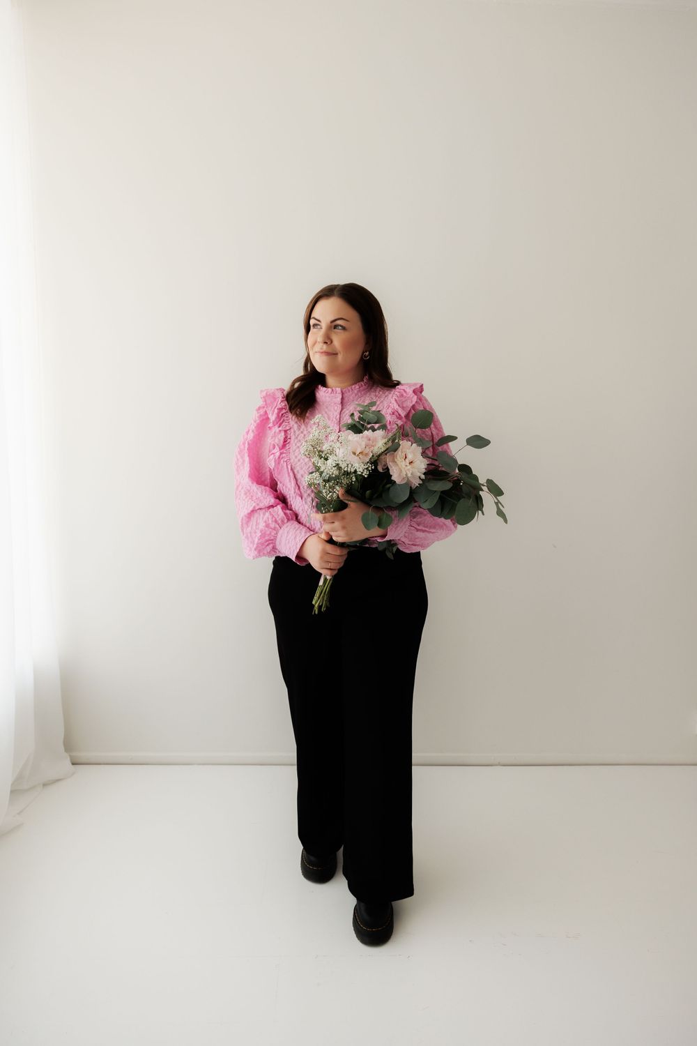 Person stands against white wall wearing pink sweater and black pants while holding bouquet of white flowers.