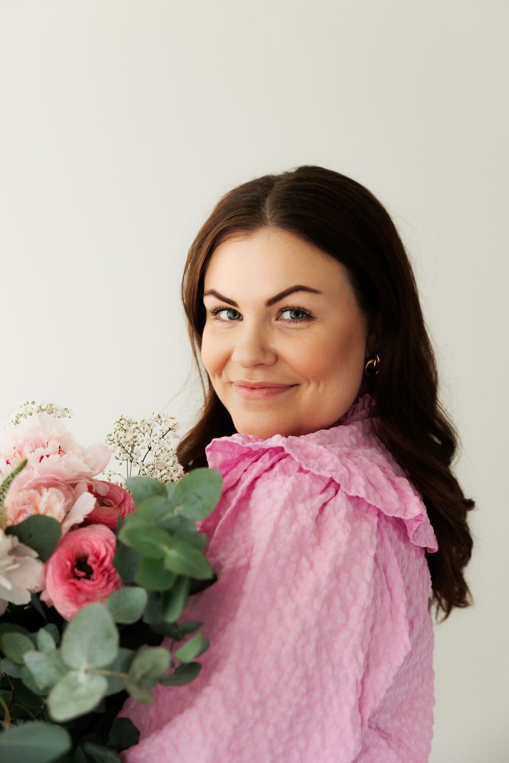 Portrait against light background wearing pink ruffled blouse and holding bouquet of pink flowers with greenery.