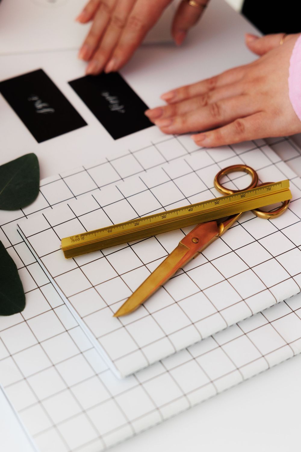 Gold measuring tools and scissors laid on white grid paper with hands arranging black business cards and green leaves.