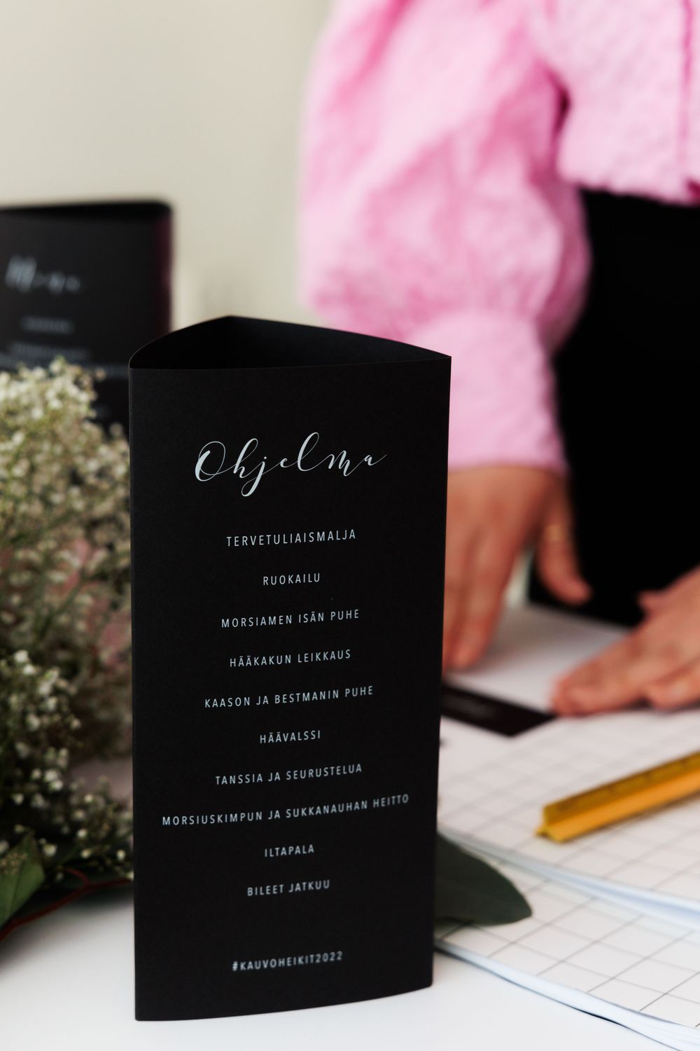 Black event menu with calligraphy text displayed on desk with baby's breath flowers and notebook.