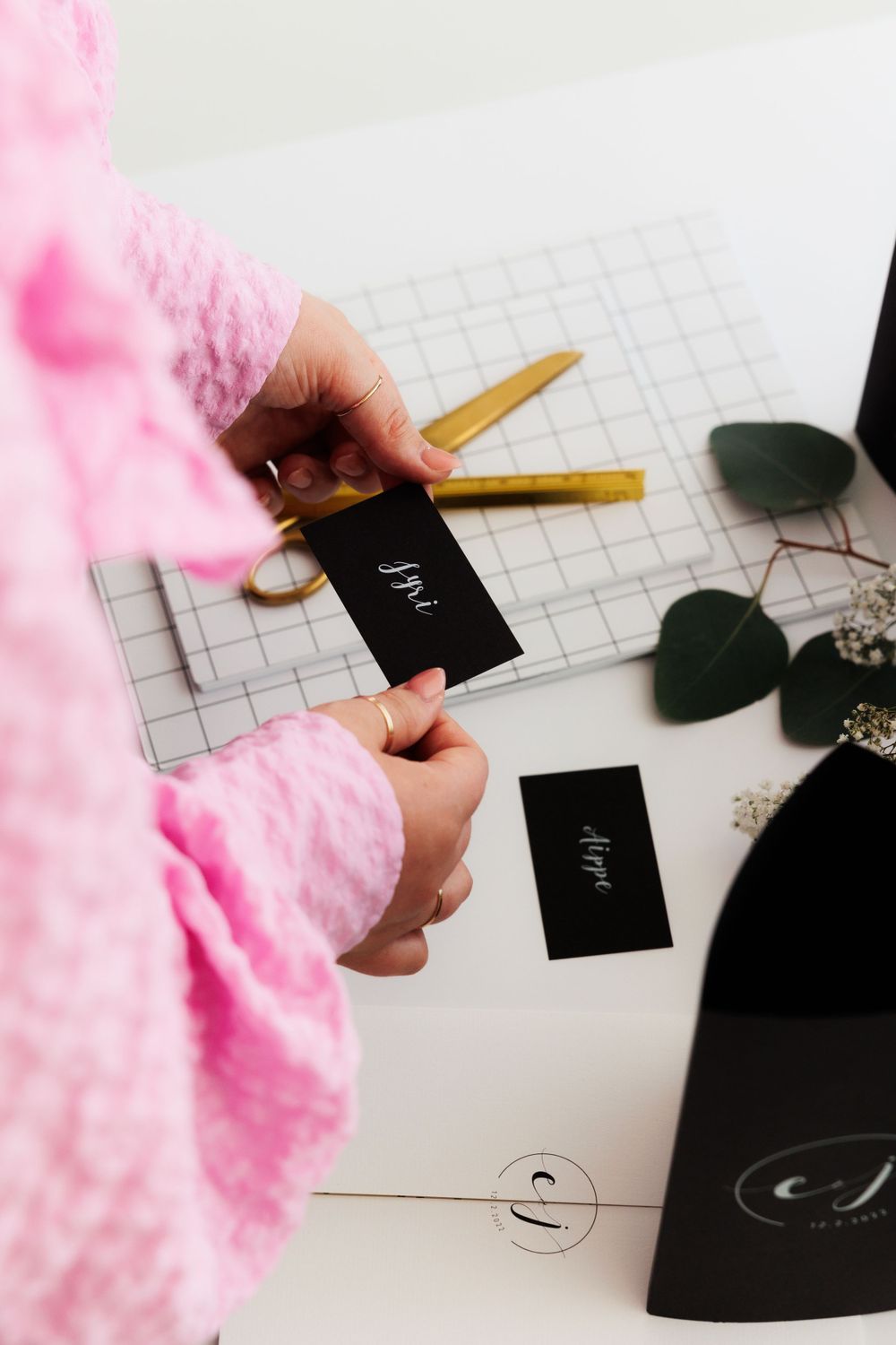 Hands in pink sleeve hold black business cards over grid paper with eucalyptus leaves and office supplies.