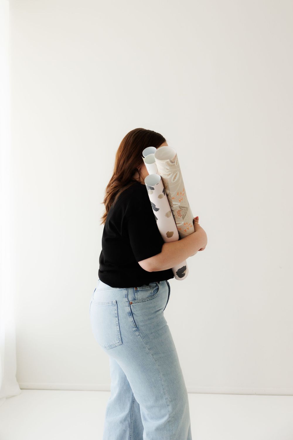 Person in black top and light jeans holding a rolled up light colored item against white wall background.