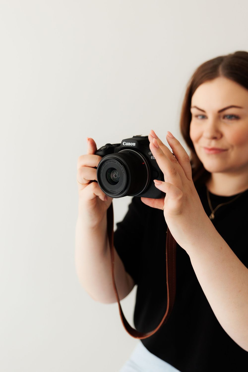 Photographer holding a Canon DSLR camera while wearing a black top against a white background.