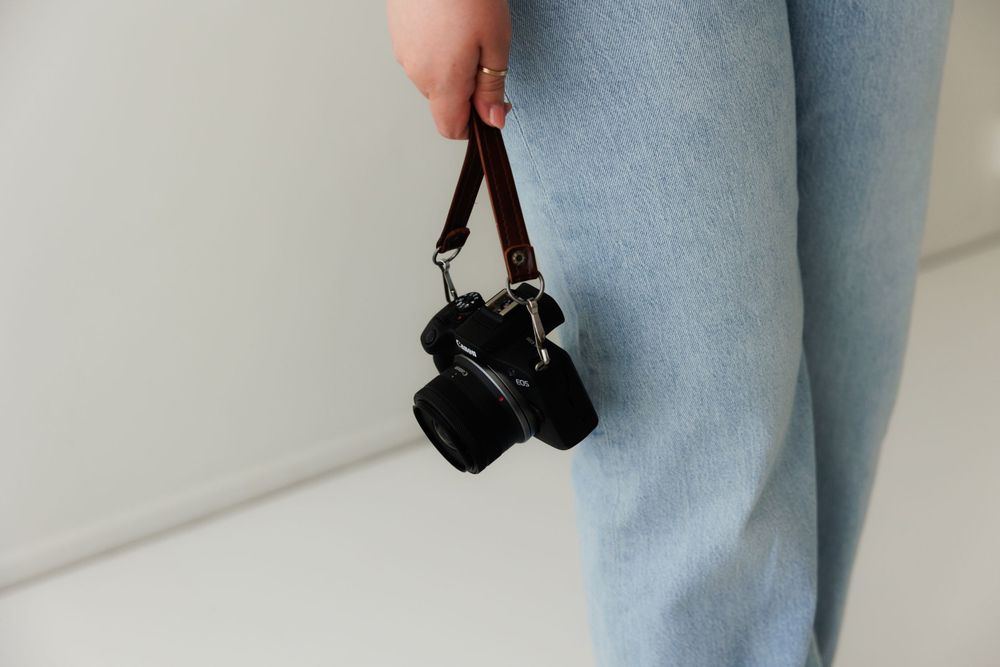 A black digital camera hangs from someone's hand wearing light denim jeans against a white wall.