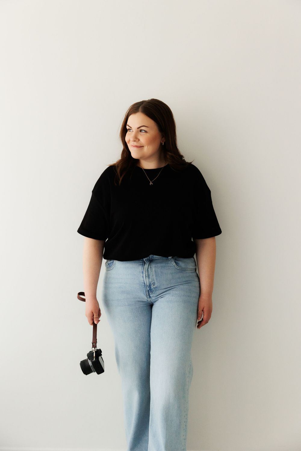 Portrait of someone in a black t-shirt and light wash jeans holding a camera against a white wall.