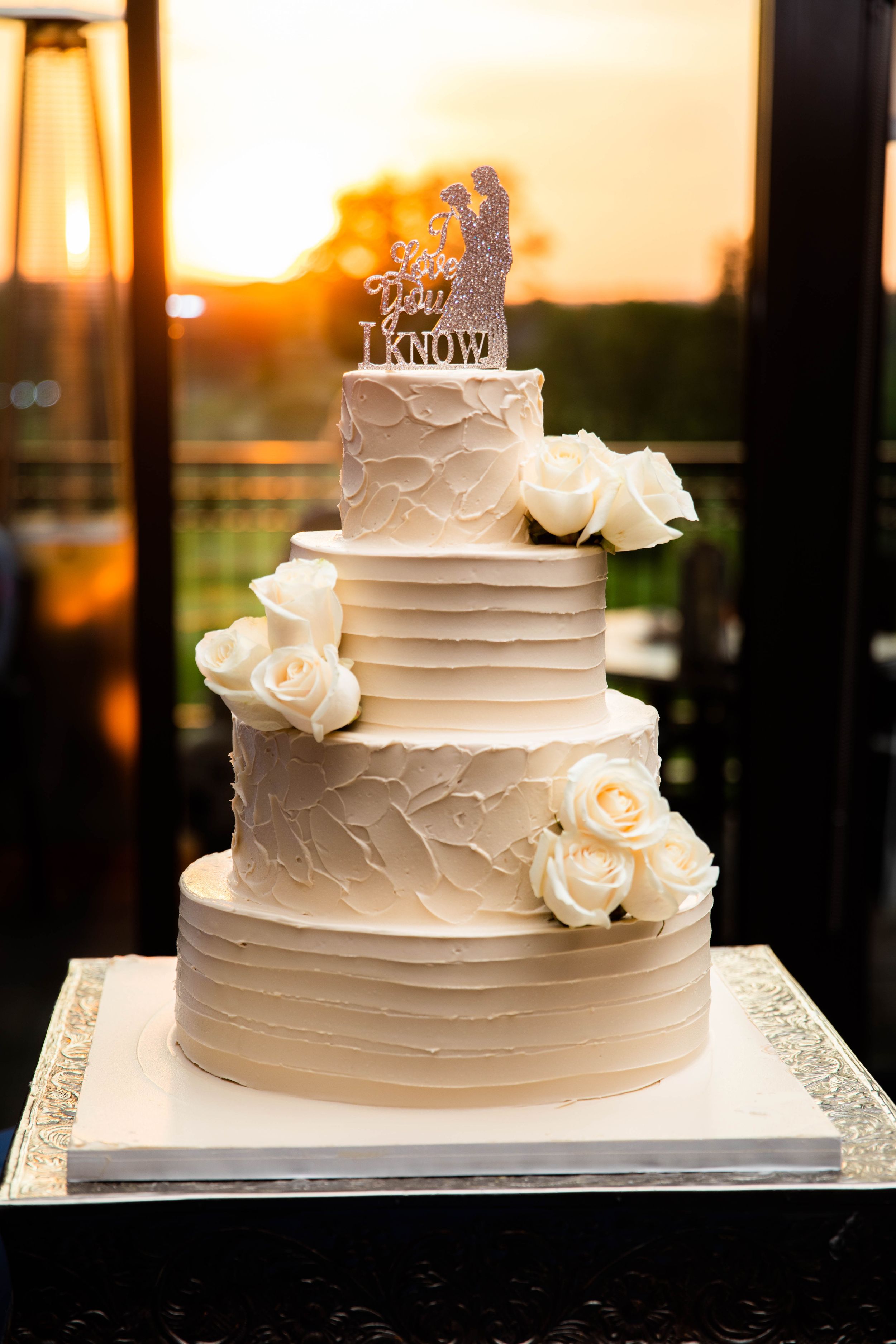 An elegant white wedding cake decorated with roses and a custom topper against a sunset backdrop.