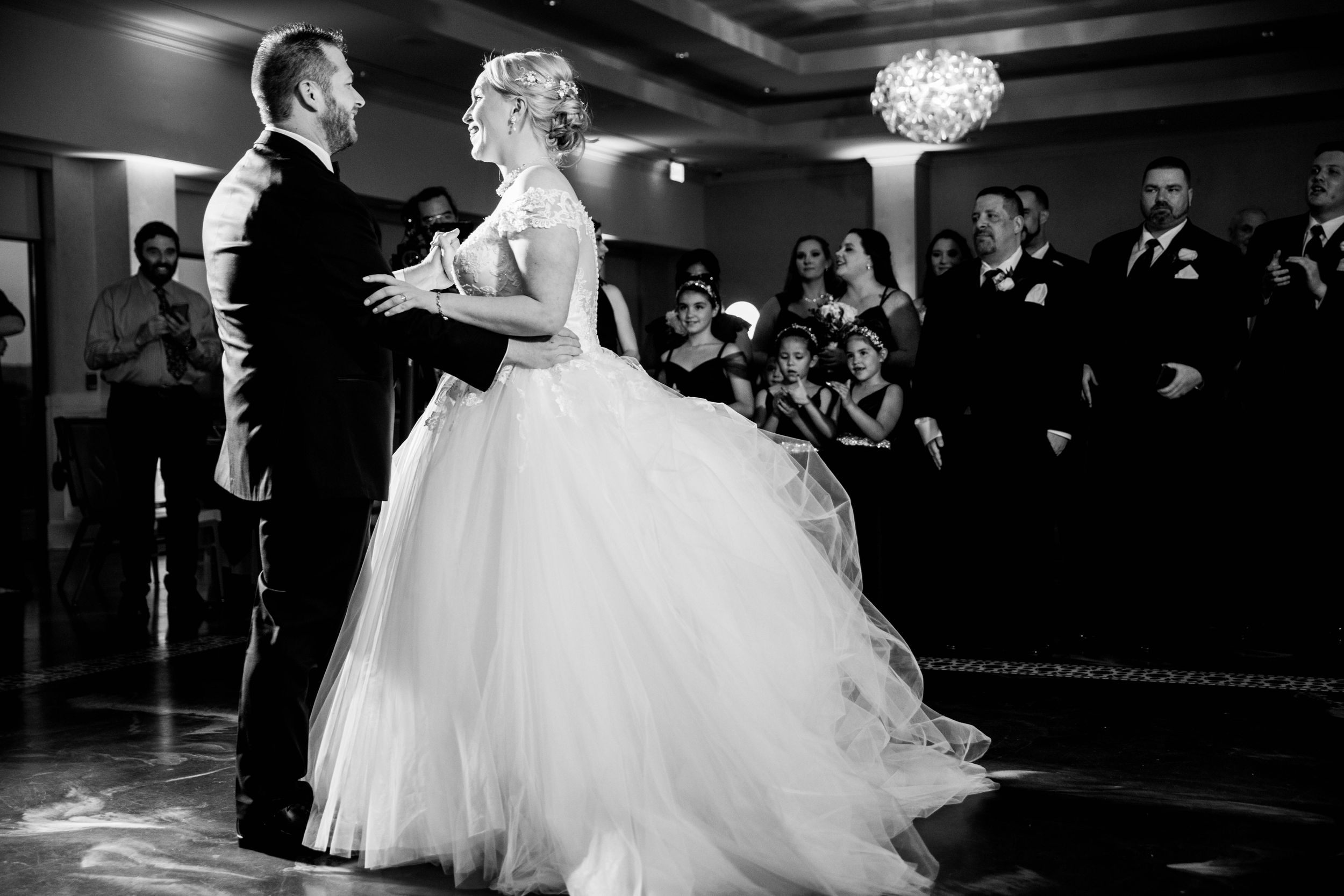 A couple shares their first dance in an elegant ballroom beneath a crystal chandelier.