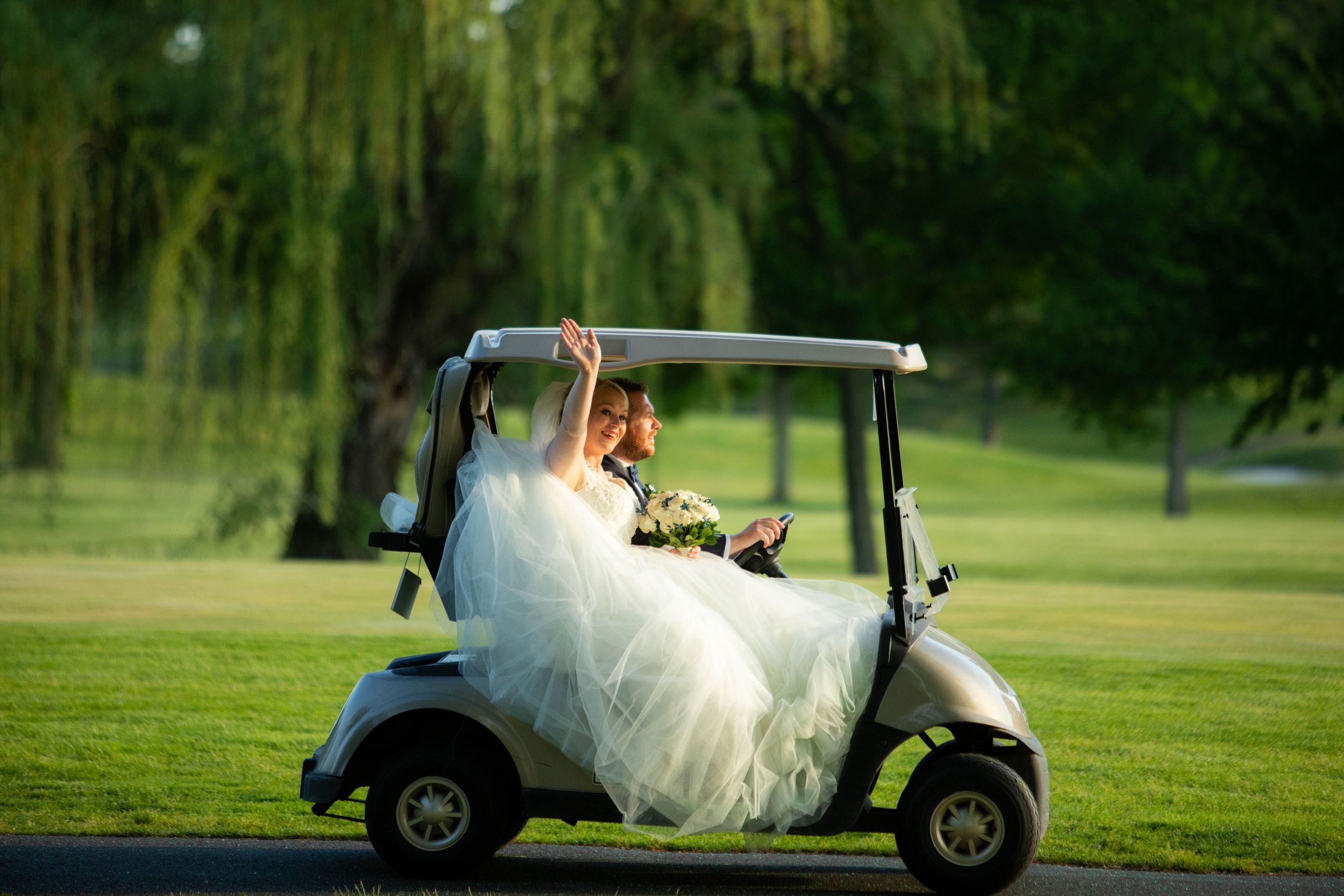 A bride in a flowing white dress sits in a golf cart on a lush green course.