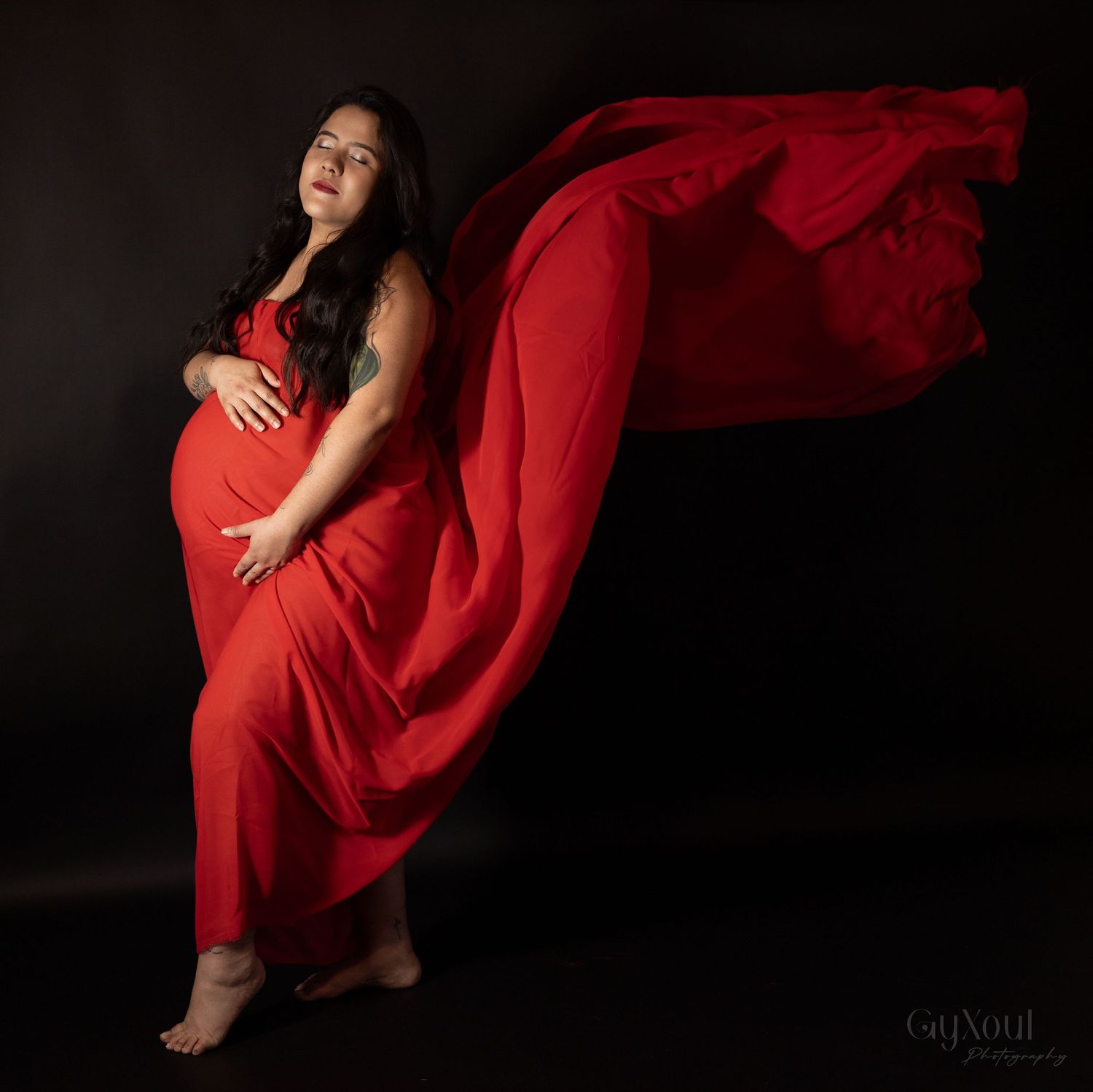 A maternity photoshoot featuring a flowing red dress against a dark background.