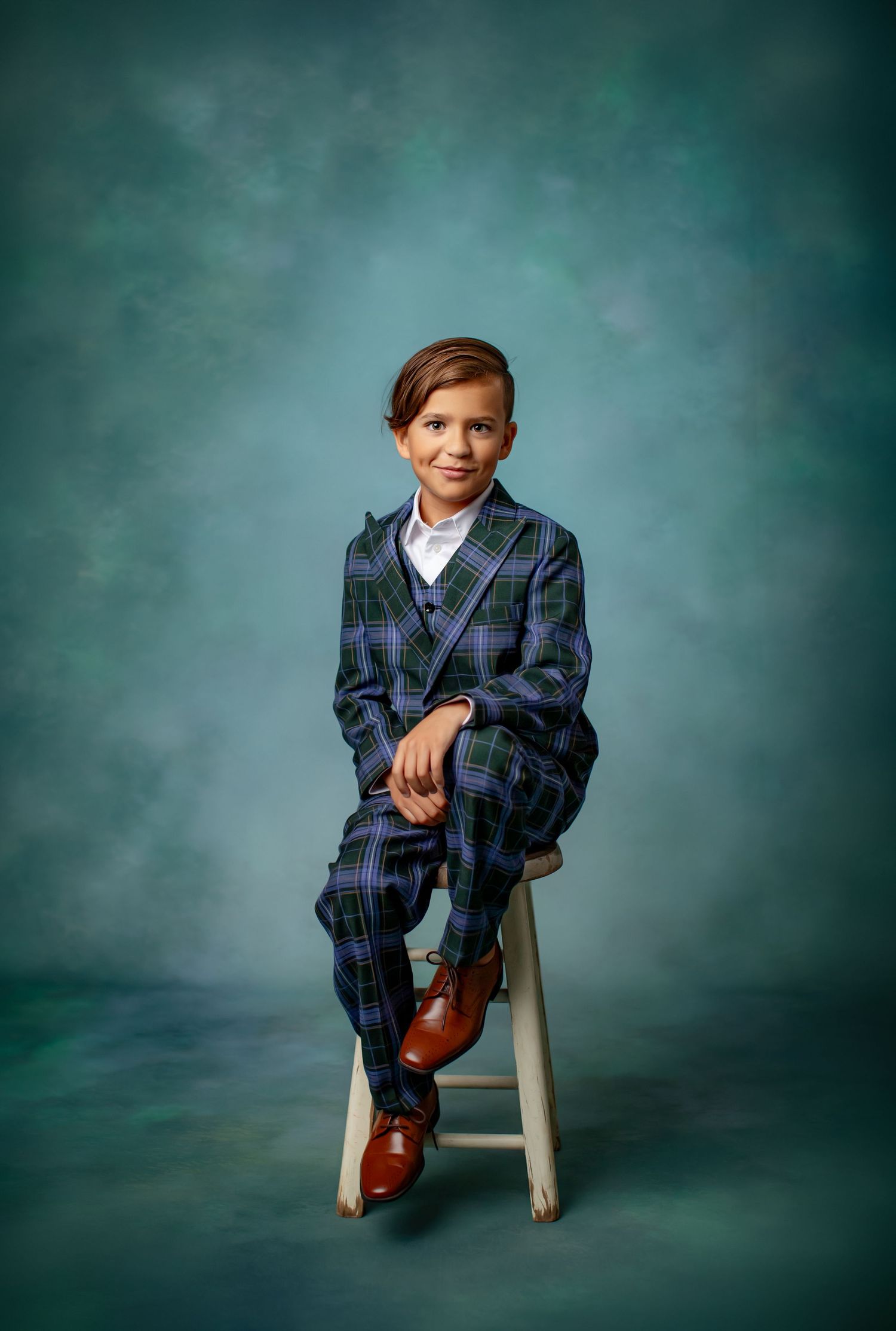 A young figure in a plaid suit sits posed on a white stool against a teal studio backdrop.