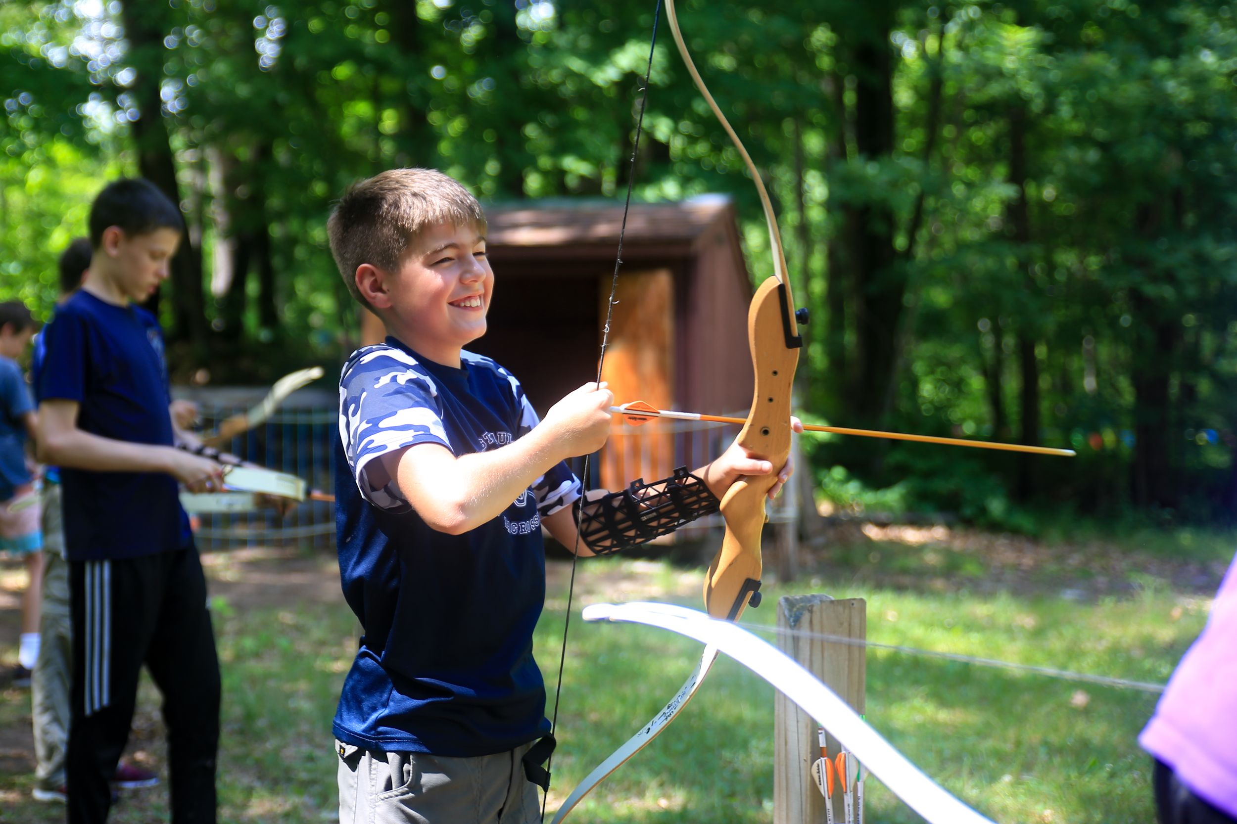 Young archer drawing a bow at an outdoor summer camp archery range in a wooded setting.