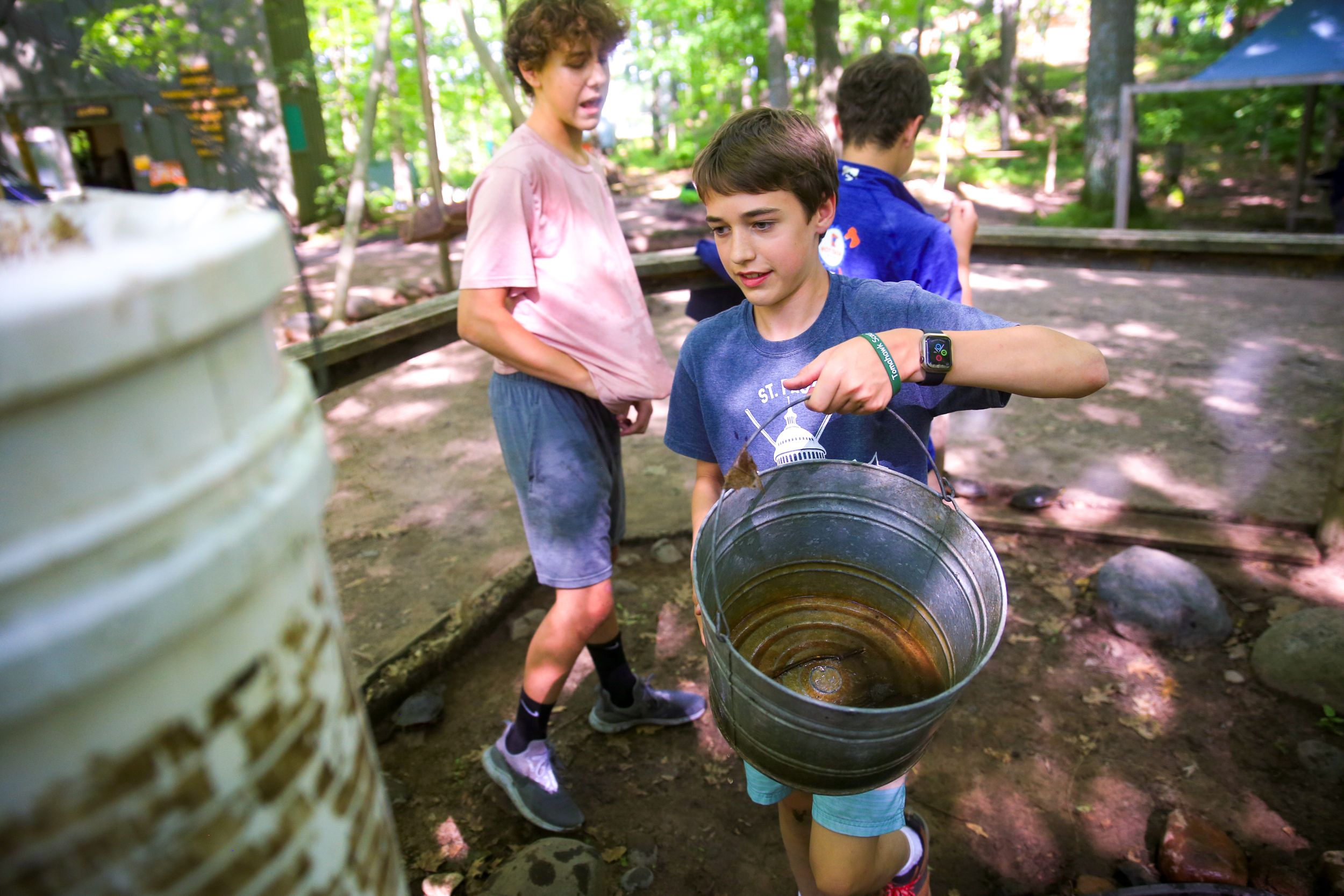 Campers examining contents of a large metal bucket in a shaded forest setting.